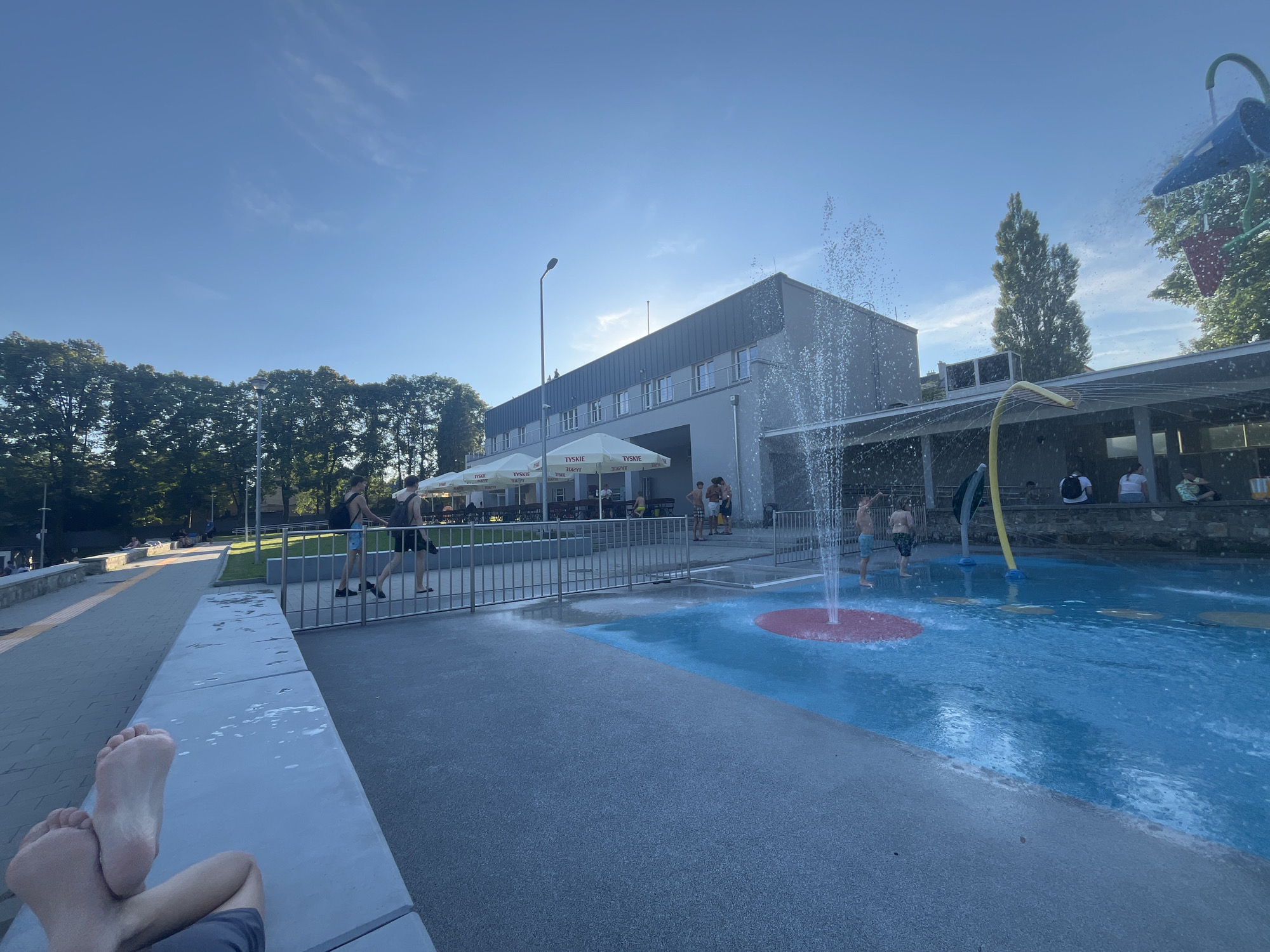 In the city of Unknown, Unknown, on a clear day with a bright blue sky, a scene unfolds at a water park. A modern, gray building with multiple windows and a dark roof stands in the background, partially shaded by white umbrellas bearing the "TYSKIE" logo. To the left of the building, a row of lush green trees borders the scene.
In the foreground, a person's bare feet and legs are visible, resting on a light gray, concrete ledge. Beyond this ledge, a splash pad area is in full operation, with water spraying from various points. The splash pad features bright blue water and a red circular section on the ground. Several children and adults are scattered around the splash pad, some interacting with the water features.
A metal fence separates the splash pad from a paved walkway. Two young men are walking along this walkway, one in a black tank top and the other in a black t-shirt, both carrying backpacks. Further down the walkway, other individuals are seated or walking, enjoying the outdoor environment. To the right of the building, a tall, slender tree stands out against the sky. The overall impression is a lively and refreshing summer day at a recreational facility.