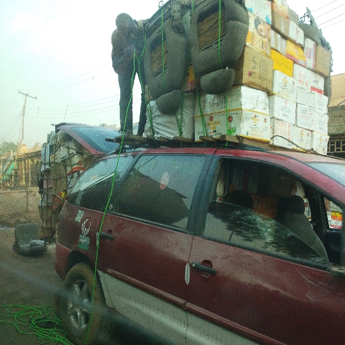 A maroon minivan is overloaded with boxes and other items on the roof. A person in dark clothing stands atop the load, securing it with rope. Additional cargo is piled high in the open hatchback area of the vehicle. The scene appears to be on a road or street with buildings and power lines visible in the background. The image is somewhat hazy and the overall impression suggests a situation of precarious and excessive loading.