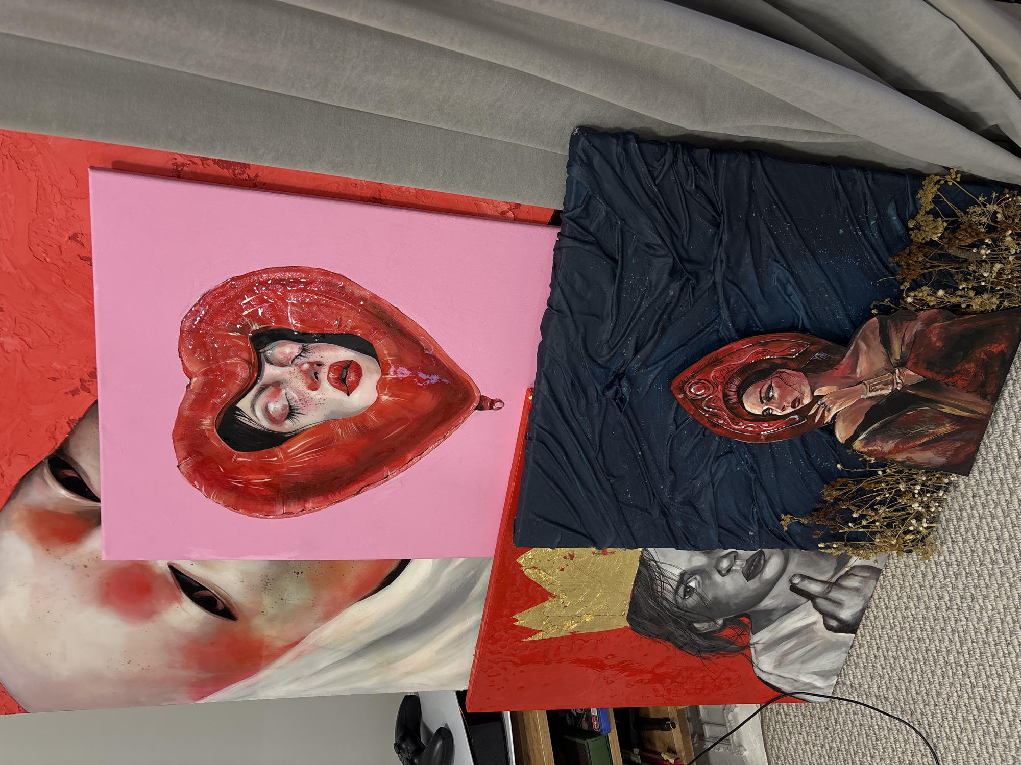 Multiple contemporary artworks are stacked and displayed in what appears to be an indoor setting in Krasnoyarsk, Russia. The arrangement shows four distinct paintings, primarily portraits, against a background of a grey pleated fabric at the top and a grey carpeted floor at the bottom.
One prominent painting features a stylized female face with closed eyes and red lips, encapsulated within a glossy red heart-shaped balloon, set against a vibrant pink background. Another painting, situated to the right, depicts a figure with a red hood or hat, lying down against a dark blue, textured background that resembles draped fabric. Dried flowers are incorporated into the bottom right corner of this piece, adding a three-dimensional element.
Partially visible at the bottom left is a large-scale portrait of a face with prominent red accents and dark, intense eyes. Below the dark blue painting, another artwork shows a grayscale portrait of a person making a defiant middle finger gesture, adorned with a golden, textured element resembling a crown, all set on a vivid red textured background.
The artworks exhibit a mix of surreal and expressive portraiture, characterized by bold colors, textural elements, and distinct emotional tones. Objects like a white console (possibly a PlayStation) and a black cable are visible on the carpeted floor, suggesting a casual, lived-in environment. There is no visible text in the image.