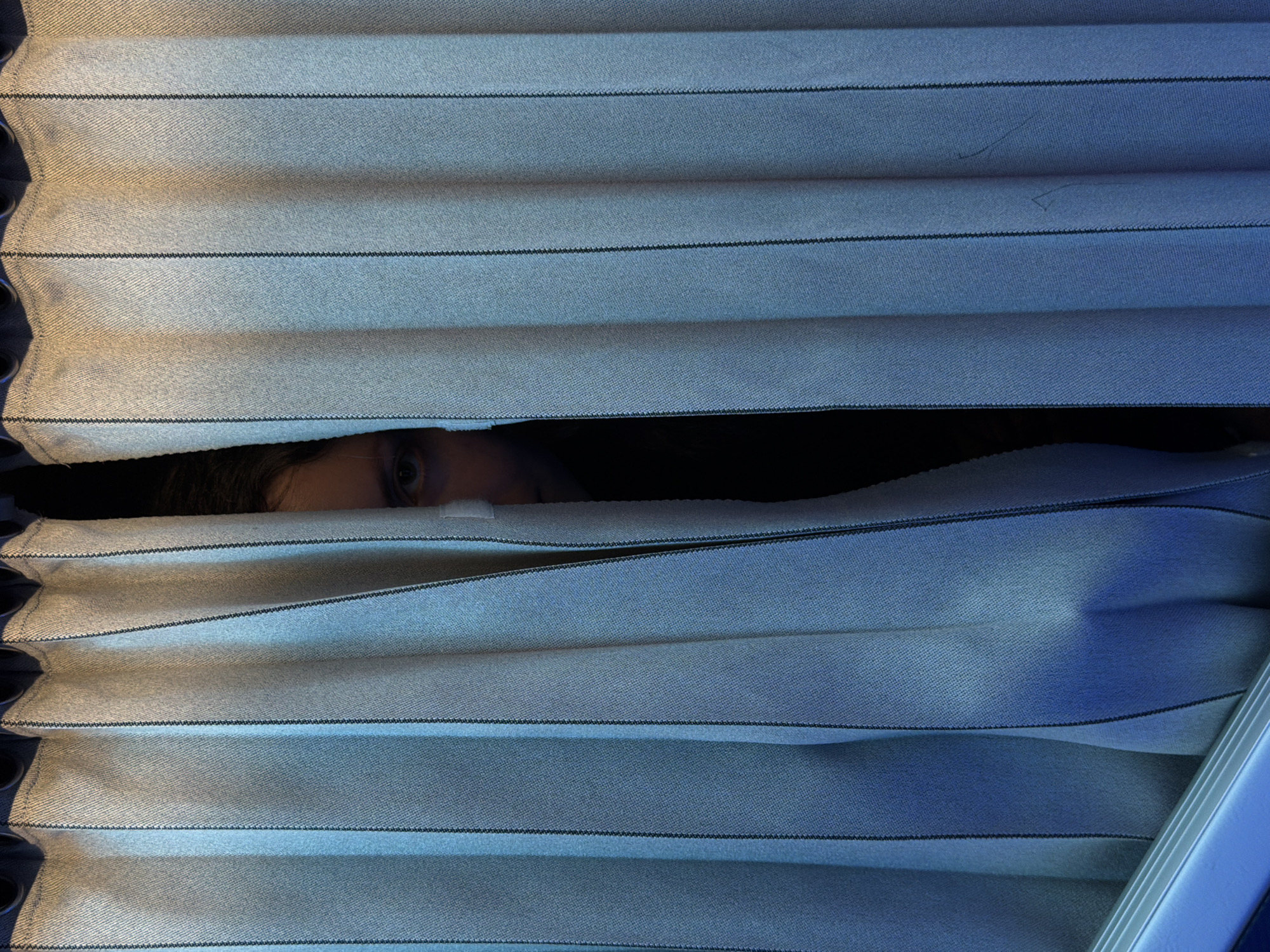 The image presents a close-up, horizontal view of a person partially obscured by pleated blinds. The blinds are light-colored, possibly off-white or light grey, with thin black horizontal stripes that appear to be stitching or decorative lines. The fabric of the blinds shows a distinct woven texture.
A single eye, part of the forehead, and the bridge of the nose of a person are visible in a narrow gap between two of the horizontal folds of the blinds, suggesting the person is peeking through. The area immediately behind the person, where the rest of their face and head would be, is dark.
The lighting varies across the blinds; the left side appears to have a warmer, slightly yellowish tone, while the right side is distinctly cast in cool blue light, hinting at an external light source or ambient lighting conditions. The visible eye appears focused, conveying a sense of observation, curiosity, or perhaps caution.
The scene depicts an act of discreet observation or looking out from a confined or hidden position. There are no specific visual cues within the image to indicate the location, but the context provided places it in Hà Nội, Vietnam. No text is visible in the image.