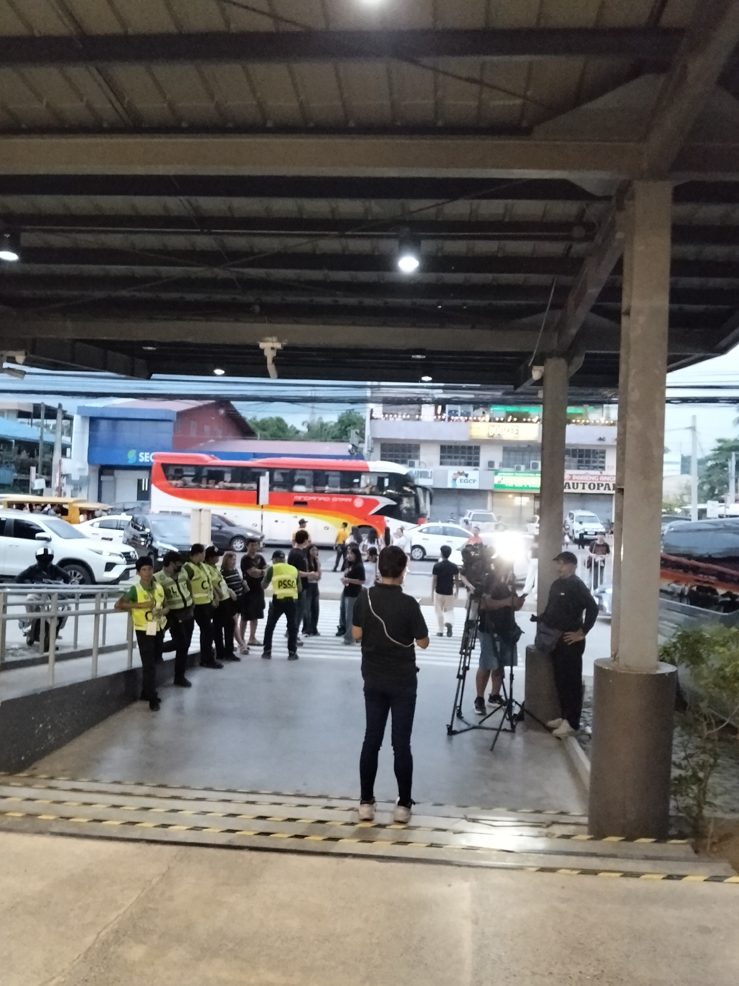 The image depicts an outdoor scene in what appears to be a transportation terminal or a busy street in Davao City, Philippines. A large, red and white bus, possibly a provincial bus, is parked in the background, alongside other vehicles including white SUVs and a tricycle. A group of individuals wearing yellow vests with "PSSC" printed on them are standing in the middle of the frame, some appearing to be security personnel or marshals. A camera crew, with one person operating a video camera on a tripod and another holding a microphone, is actively filming the group in yellow vests. Several other people are scattered throughout the scene, some observing the activity and others going about their business. The environment suggests a public space with a covered walkway or structure overhead, supported by pillars. The lighting indicates it is either dusk or early morning. The overall impression is that of a news reporting or event coverage scenario taking place in a public area of Davao City.