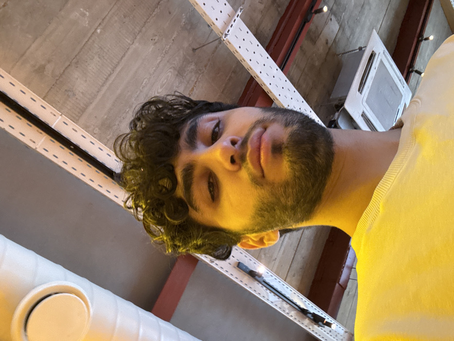 A selfie captures a man with curly dark hair and a beard looking down at the camera. He wears a yellow shirt. The perspective is from below, showing the ceiling with white and red structural elements. An air conditioning unit is visible in the upper right corner. The lighting gives a warm, slightly yellow tone to the image.