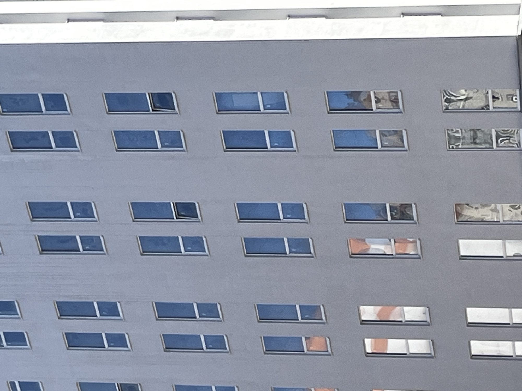 This media file displays a close-up, eye-level view of the exterior of a modern building, likely an apartment complex or office building, in Saint Petersburg, Russia. The facade is a uniform light gray, punctuated by rows of horizontal, rectangular windows. The windows are divided into two panes, with a thin white frame separating them.
The windows reflect the environment. Some show a clear blue sky, suggesting it is daytime. Others reflect the surrounding architecture and possibly the sky, with distorted patterns of light and shadow. A few windows appear to be partially open, hinting at a mild or warm day. The light indicates it is likely daytime, possibly mid-morning or afternoon, with the sun casting some reflections.
The overall impression is one of urban architecture and the everyday scene of a building with multiple dwelling or office units. There is no discernible activity of people, but the open windows suggest occupancy. The building appears to be tall, given the perspective, and its repetitive design is characteristic of modern urban construction. No text is visible in the image. The weather appears to be clear and sunny.