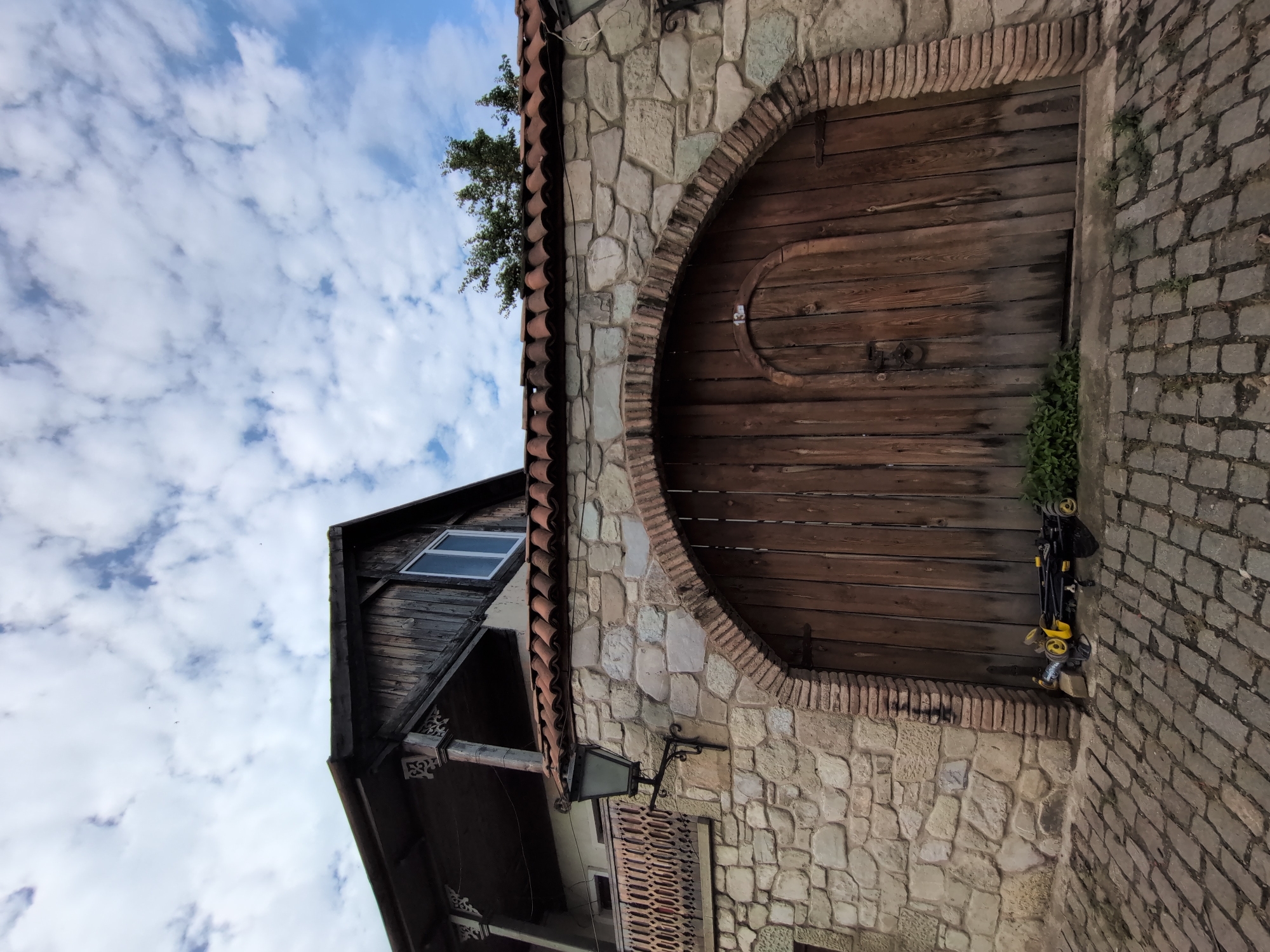 The image shows a weathered wooden gate set into a stone wall in Mtskheta, Georgia. The gate is arched and has a smaller, arched door integrated into its design. A metal ring serves as a handle for the main gate. To the left of the gate, leaning against the stone wall, is a yellow and black foldable stroller. Greenery grows along the base of the gate and wall. Above the gate, a traditional tiled roof tops the stone structure, and a wooden balcony with decorative railings extends from the building behind the wall. A black wrought-iron lantern is mounted on the stone wall. The ground in front of the gate is paved with cobblestones. The sky above is partly cloudy with patches of blue.