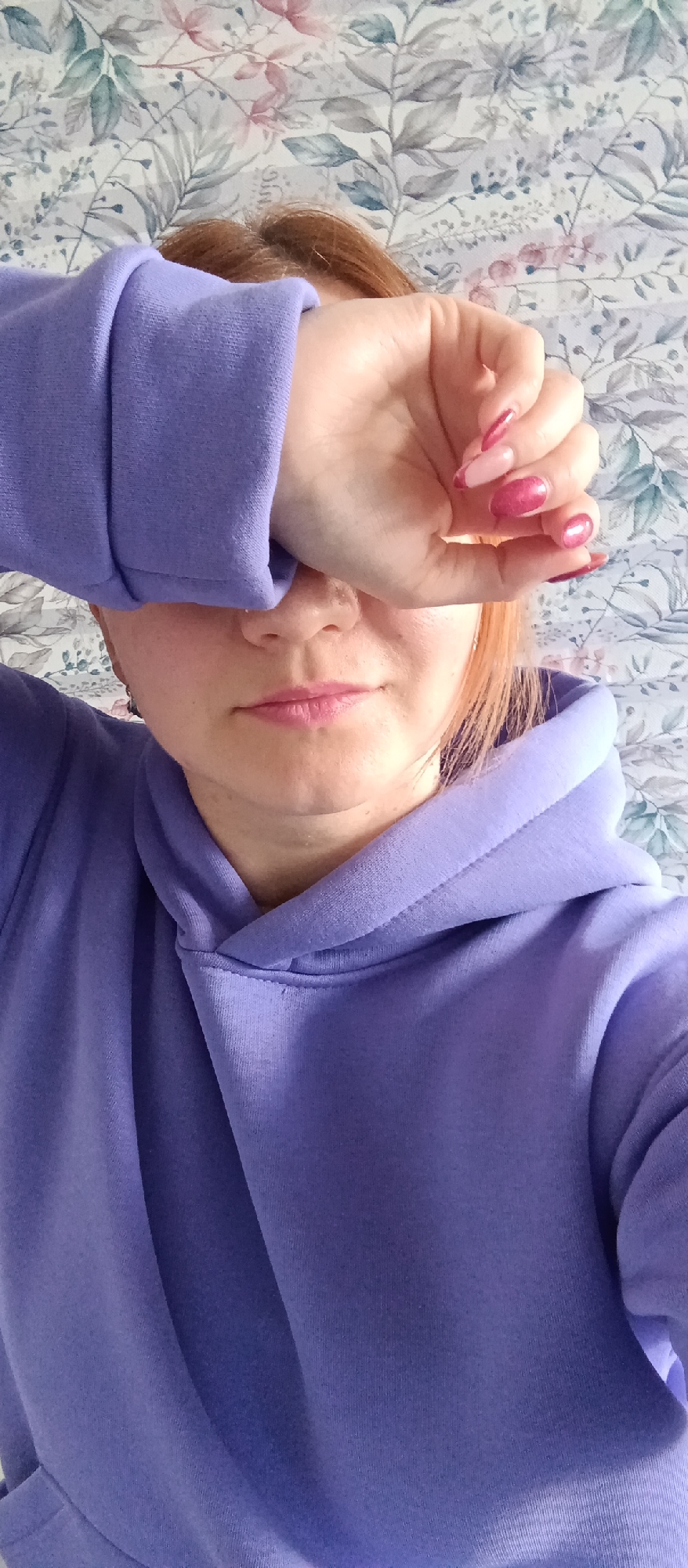 A person wearing a purple hoodie obscures their eyes with their hand. The person has reddish-blonde hair, and their nails are painted pink with a glittery finish. The background is a floral-patterned wallpaper. The location is unknown.