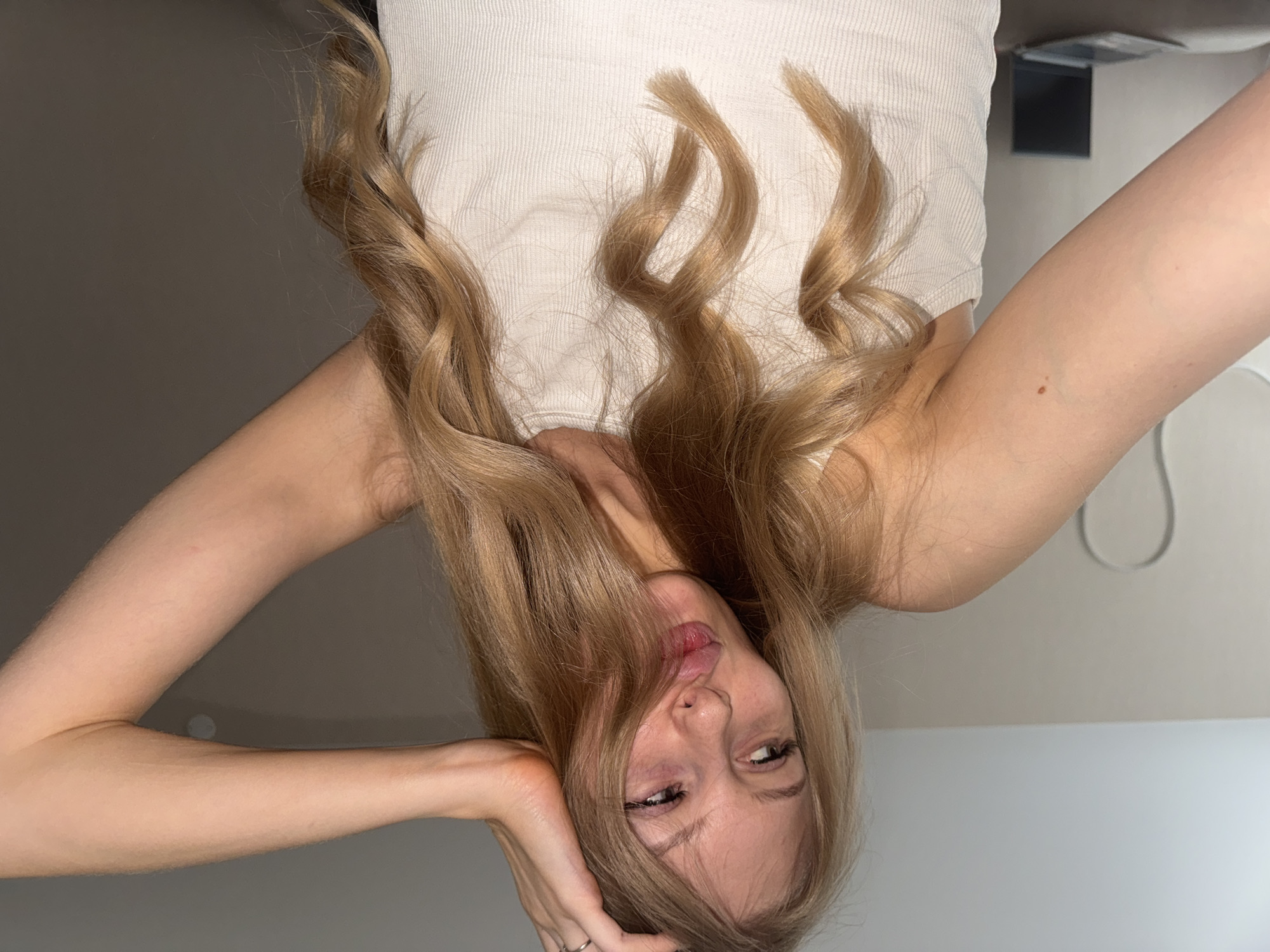 A fair-skinned woman with light blonde hair is posing for a photograph. She is upside down, resting her head on her left hand and looking slightly to her right. She is wearing a light-colored tank top. The background consists of neutral-colored walls and part of a light fixture. There is no information to indicate this photograph was taken in Perm, Russia.