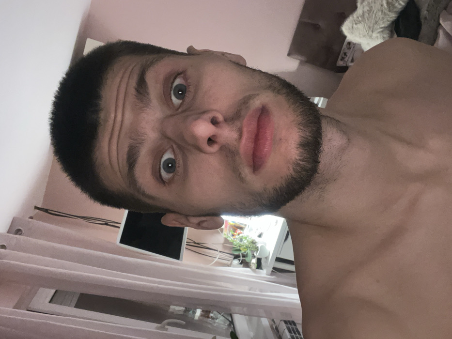 The image displays a close-up, frontal portrait of an adult male situated indoors in Karan Varbovka, Bulgaria. The subject, estimated to be in his late twenties to early thirties, possesses short dark hair and a short beard. He is bare-chested, revealing his shoulders and upper chest. His facial expression includes wide-open eyes and slightly pursed lips, directed towards the camera. The background features a light pink wall. To the subject's left, light-colored curtains with visible grommets partially cover a window, allowing ambient light to enter. A dark-screened monitor or television is mounted or placed on a surface in the mid-ground, with multiple dark cables visible behind it. Below this, a white desk or shelf holds several small potted plants, illuminated by an artificial light source. To the right, a section of dark furniture, possibly a headboard or chair, is visible, adorned with a light-colored, possibly faux-fur, item. The environment suggests a residential interior.