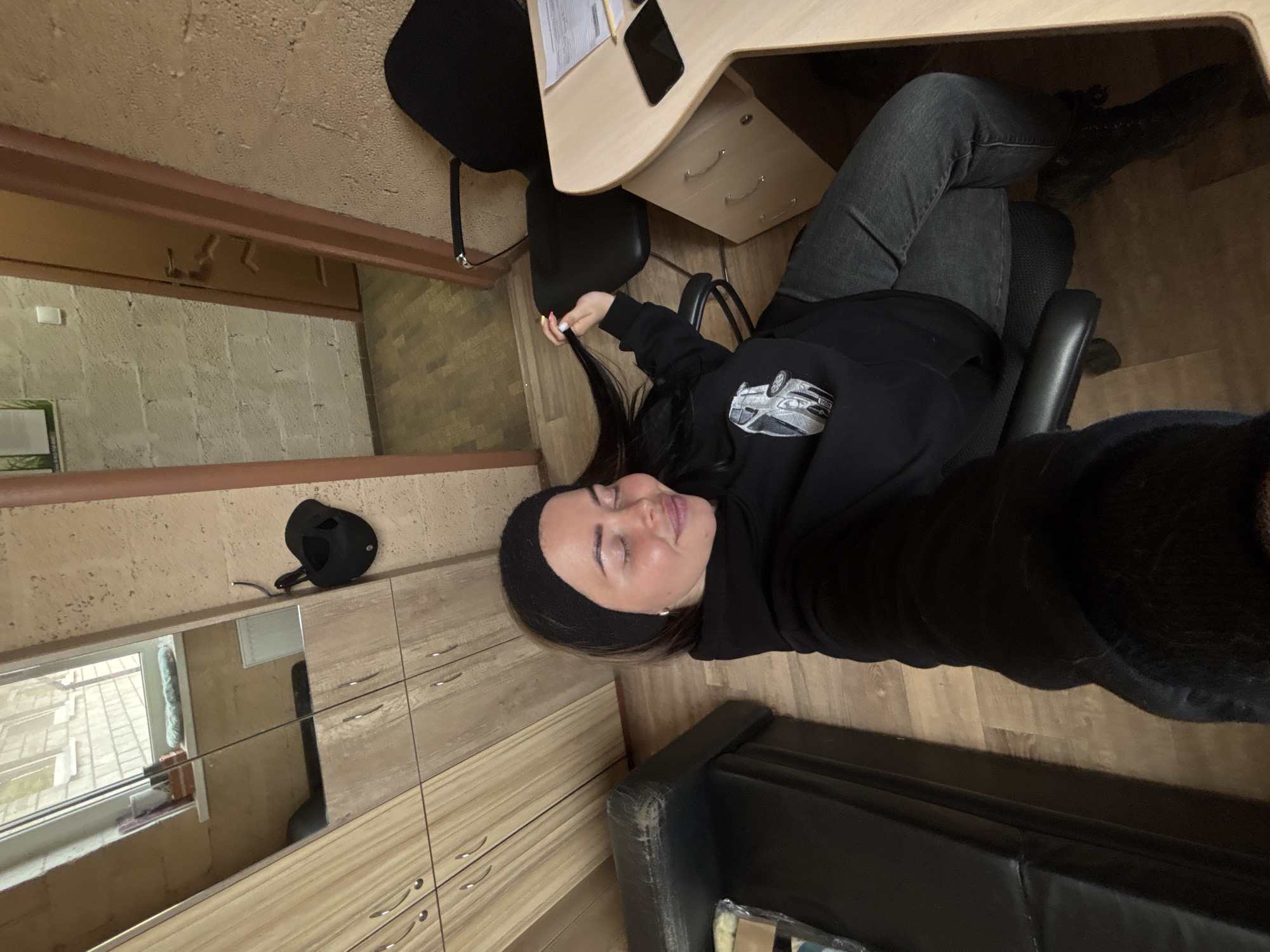 A woman with fair skin and dark hair is sitting in a black office chair at a light-colored desk. She is wearing a black hoodie with a car graphic on the front and grey jeans. She is indoors, likely in a room with light-colored walls and wood-grain laminate flooring. A black baseball cap is mounted on the wall near a mirror. A portion of a black leather couch is visible in the lower center of the image. The location is Русский Вожой, Russia.