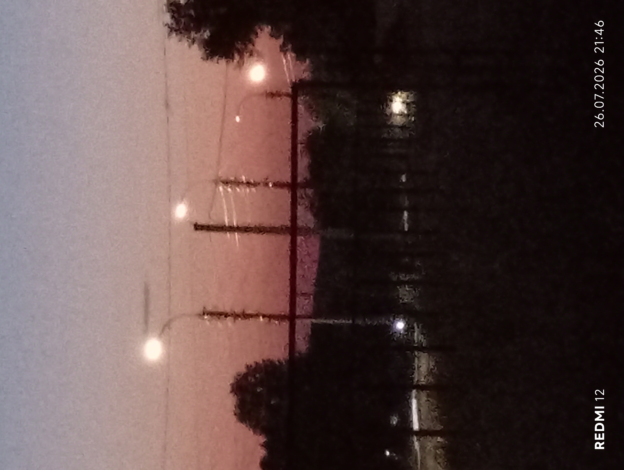 The image shows a street scene at dusk in Smila, Ukraine. The sky is a gradient of pale blue to pink. Several streetlights are illuminated, casting a glow. In the foreground, there is a dark, silhouetted area of trees and a fence. The ground appears to be a path or road with some faint lights visible in the distance. The bottom of the image contains text overlays: "REDMI 12" and "26.07.2026 21:46". There are no people or animals visible in the image.