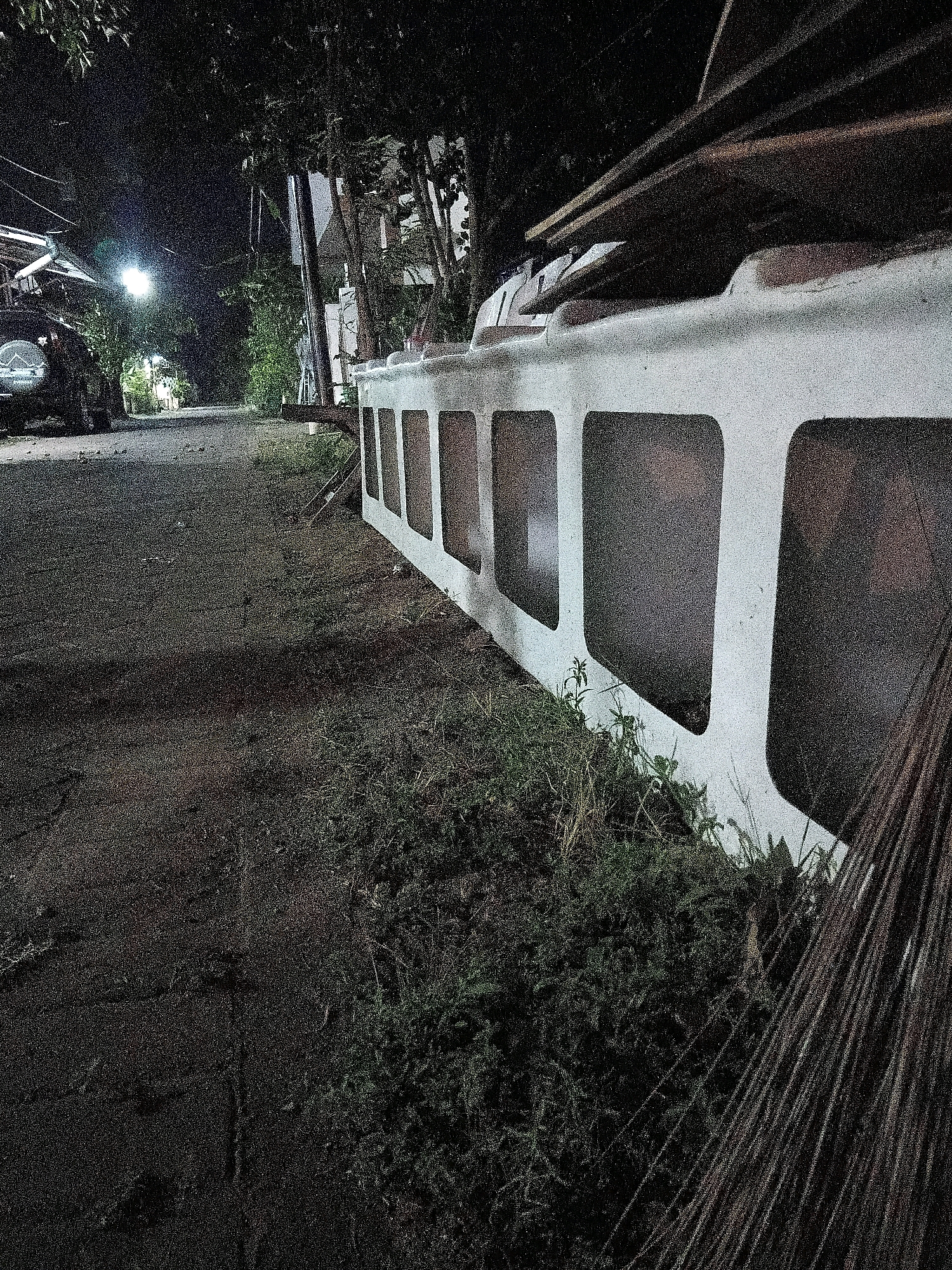 The image shows a street scene at night in Cikal, Indonesia. A white concrete barrier with rectangular openings runs along the side of the road. Several wooden planks are stacked on top of the barrier. To the left of the barrier, a paved road is visible, with a vehicle parked on the side. Trees and vegetation line the road, and a street light illuminates the scene in the background. A bundle of palm fronds lies on the ground to the right of the barrier. The overall impression is of a quiet, possibly rural or suburban area at night.