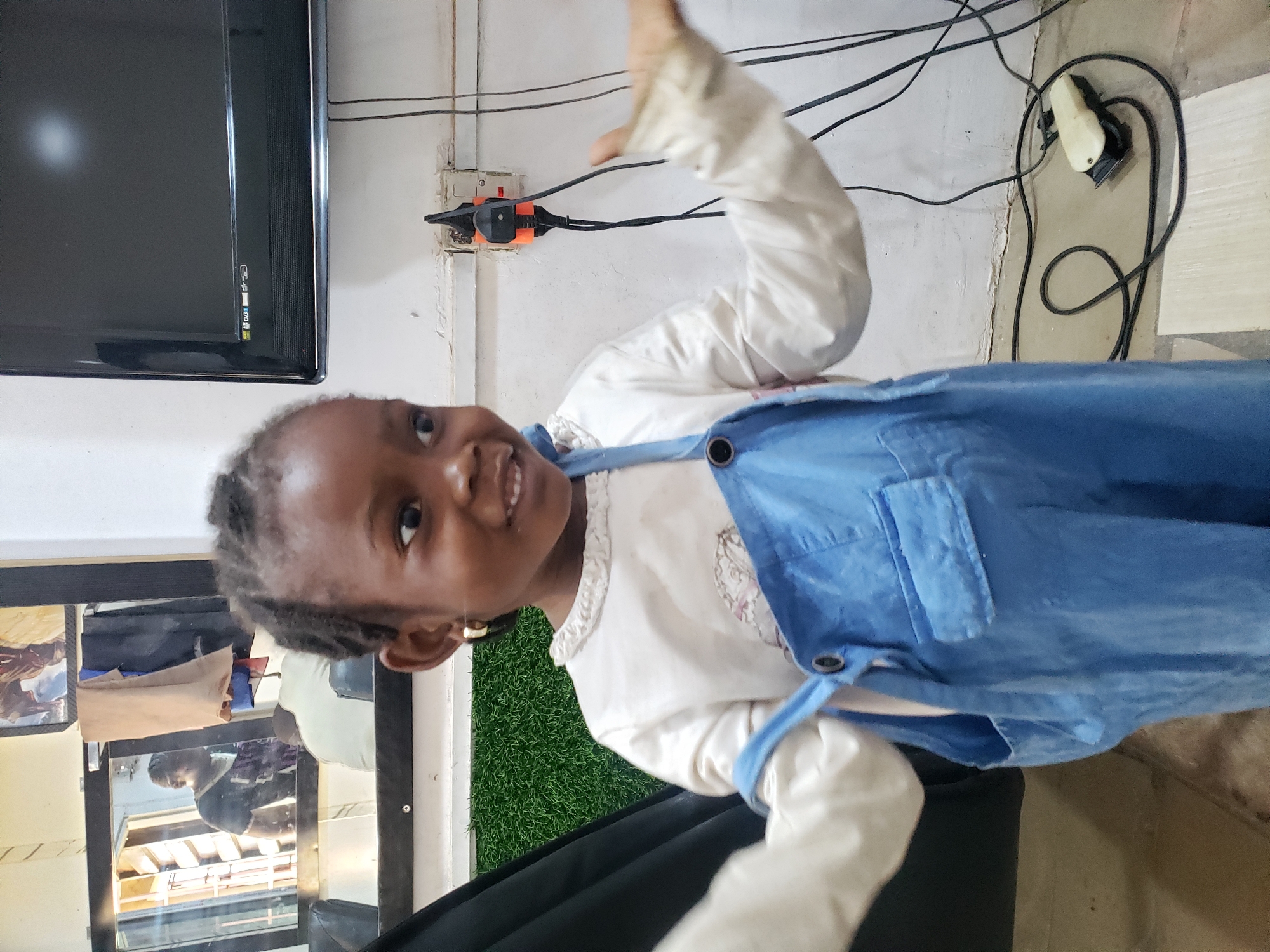 A young child, likely a girl, is featured prominently in a candid indoor shot. She has dark skin, braided hair, and is wearing a white long-sleeved top under a blue overall dress (dungarees). She is smiling broadly and looking directly at the camera, with her right arm raised in a playful or greeting gesture.
The setting appears to be a functional room, possibly a local hair salon or a multi-purpose space in Adankolo, Nigeria. To her upper left (when oriented correctly), a dark flat-screen television is mounted on a white wall, displaying a dark screen with faint icons, possibly including text like "D.J." On the wall to her right, an electrical outlet with a multi-plug adapter is visible, surrounded by surface-mounted electrical wires. Further to the right, on the floor, a white and black hair clipper with its cord is lying.
To the child's left and behind her, a large mirror reflects parts of the room and what appears to be another person or object. Adjacent to the mirror, a section of artificial green turf is visible, suggesting a decorative element or part of a salon setup. The child is sitting or standing in front of a black chair or seating area. The overall impression is one of a cheerful, spontaneous moment captured in a practical, unadorned interior space. No specific activity beyond posing is evident. The lighting suggests it is daytime.