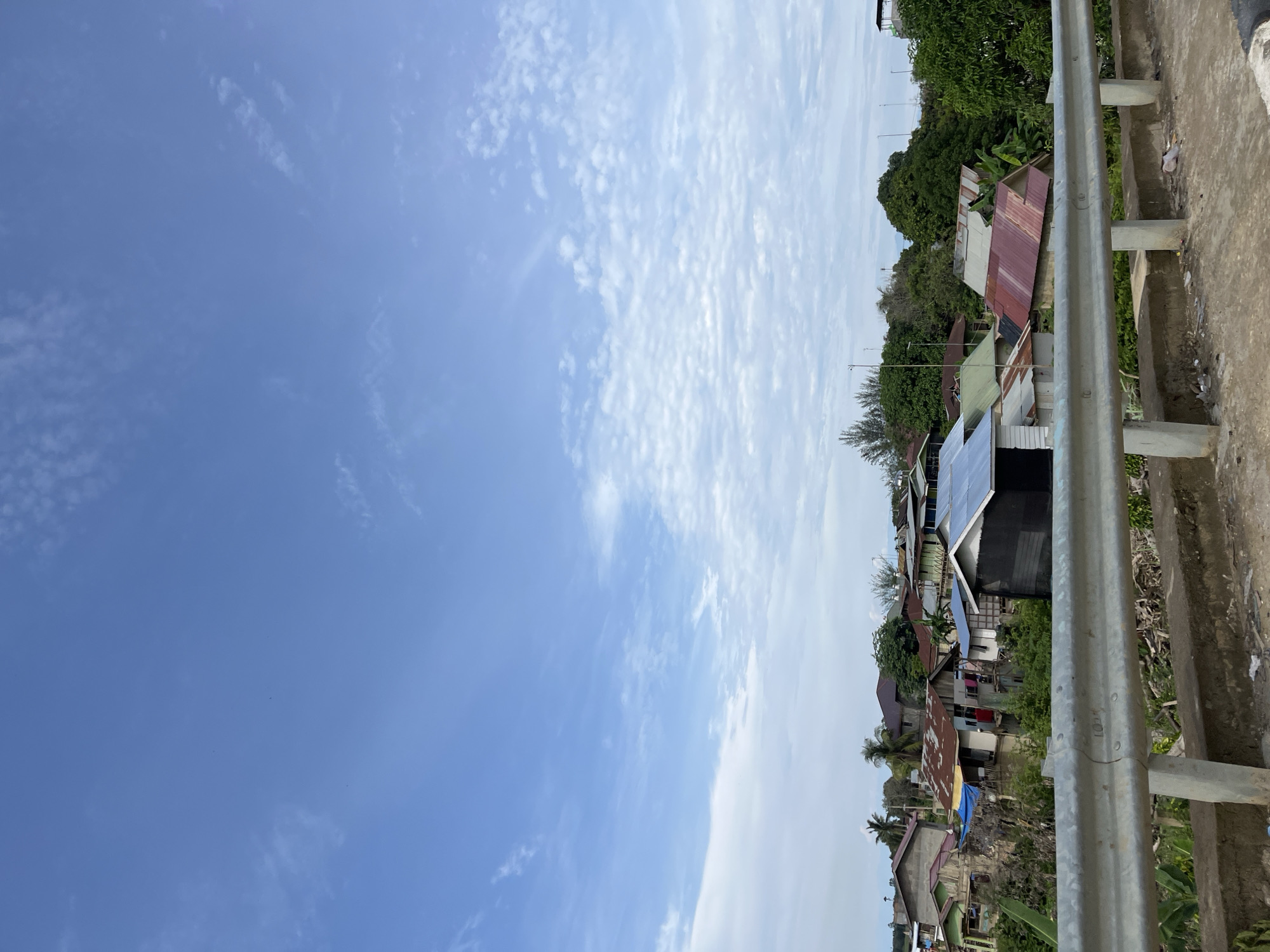 A wide shot captures a residential area in Sukajadi, Indonesia, with a metal guardrail in the foreground. The guardrail, composed of vertical concrete posts and horizontal metal bars, runs across the lower portion of the frame. Beyond the guardrail, a cluster of houses is visible, characterized by corrugated metal roofs in various colors, including red, gray, and green, and walls made of wood and concrete. Some houses appear to be elevated or situated close to water. Lush green trees and vegetation surround the houses, and palm trees are interspersed throughout the scene. The sky above is a clear blue, adorned with scattered white clouds, suggesting fair weather. The overall impression is of a densely populated, tropical urban environment.