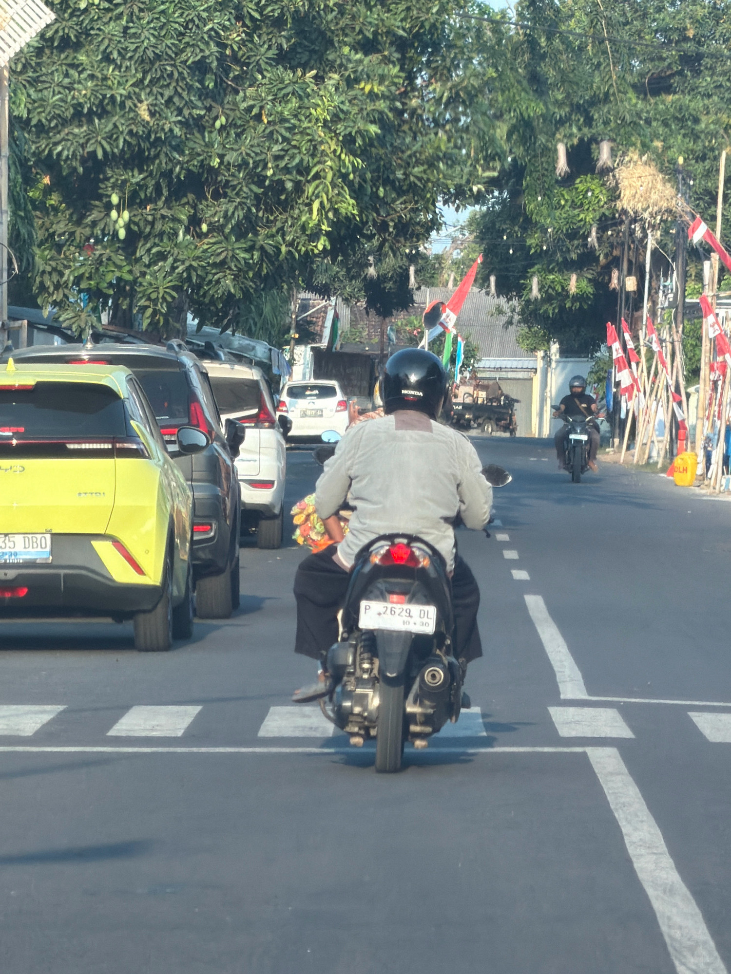 The image captures a street scene in Patokan, Indonesia. In the foreground, a person wearing a black helmet and a light-colored shirt is riding a motorcycle with a license plate "P 2629 DL". The motorcycle has a bouquet of flowers on the back. To the left, a yellow SUV with license plate "35 DBO" is stopped at a pedestrian crossing. Several other cars are lined up behind the yellow SUV. On the right side of the road, there are trees decorated with red and white flags and white streamers. Another person is riding a motorcycle further down the road, and a small truck is visible in the background. The scene appears to be a street with moderate traffic and decorative elements, possibly for a local celebration or event.