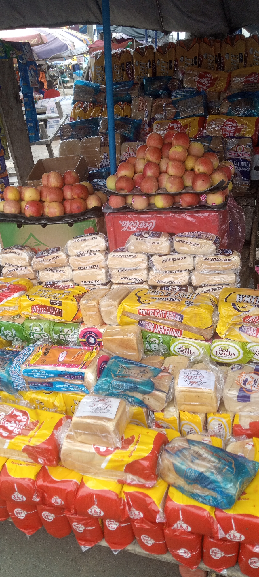 A market stall in Jalingo, Nigeria, displays a variety of baked goods and fruits. The stall is covered by a blue and grey tarp, offering shade from the sun. In the foreground, rows of bagged bread are stacked, with visible labels indicating brands like "White Light Premium Bread" and "J&J Bread". Some loaves are individually wrapped in clear plastic. To the left, a stack of apples in various shades of red and yellow are piled on top of a cardboard box. Behind the bread and apples, more bread loaves are stacked, some with visible branding such as "Blackberry Bread" and "Our Nation Super Milk Bread". A red Coca-Cola branded cooler is positioned behind the apples and some of the bread. In the background, other market stalls and shoppers can be seen. The overall scene depicts a typical marketplace environment.