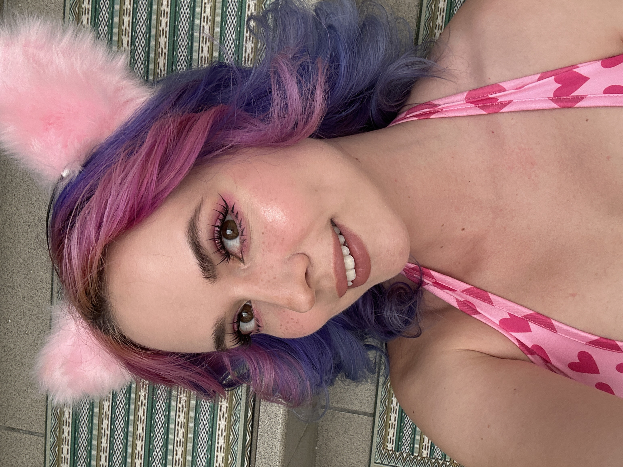 A person with pink cat ears and multi-colored hair lies on a surface with a patterned covering. The person is wearing a pink top with a heart pattern. The location is not identifiable as City of Zagreb, Croatia from the visual content.