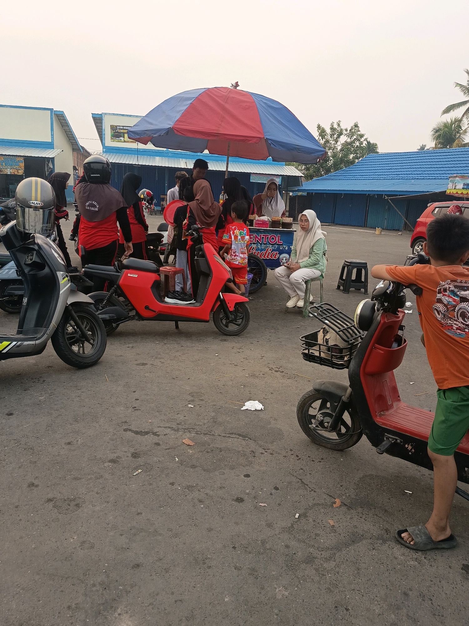 An outdoor daytime scene in Pulau, Indonesia, depicts a street with several parked electric scooters and a food stall under a large umbrella. A person wearing an orange t-shirt and green shorts stands in the foreground, leaning on a red scooter. In the background, a food stall displays food items and is attended by a person in a light green headscarf. Several other individuals, some wearing headscarves and casual clothing, are gathered around the stall and scooters. The environment appears to be a paved street or parking area, with buildings with blue roofs and white facades visible in the background. The sky is overcast.
