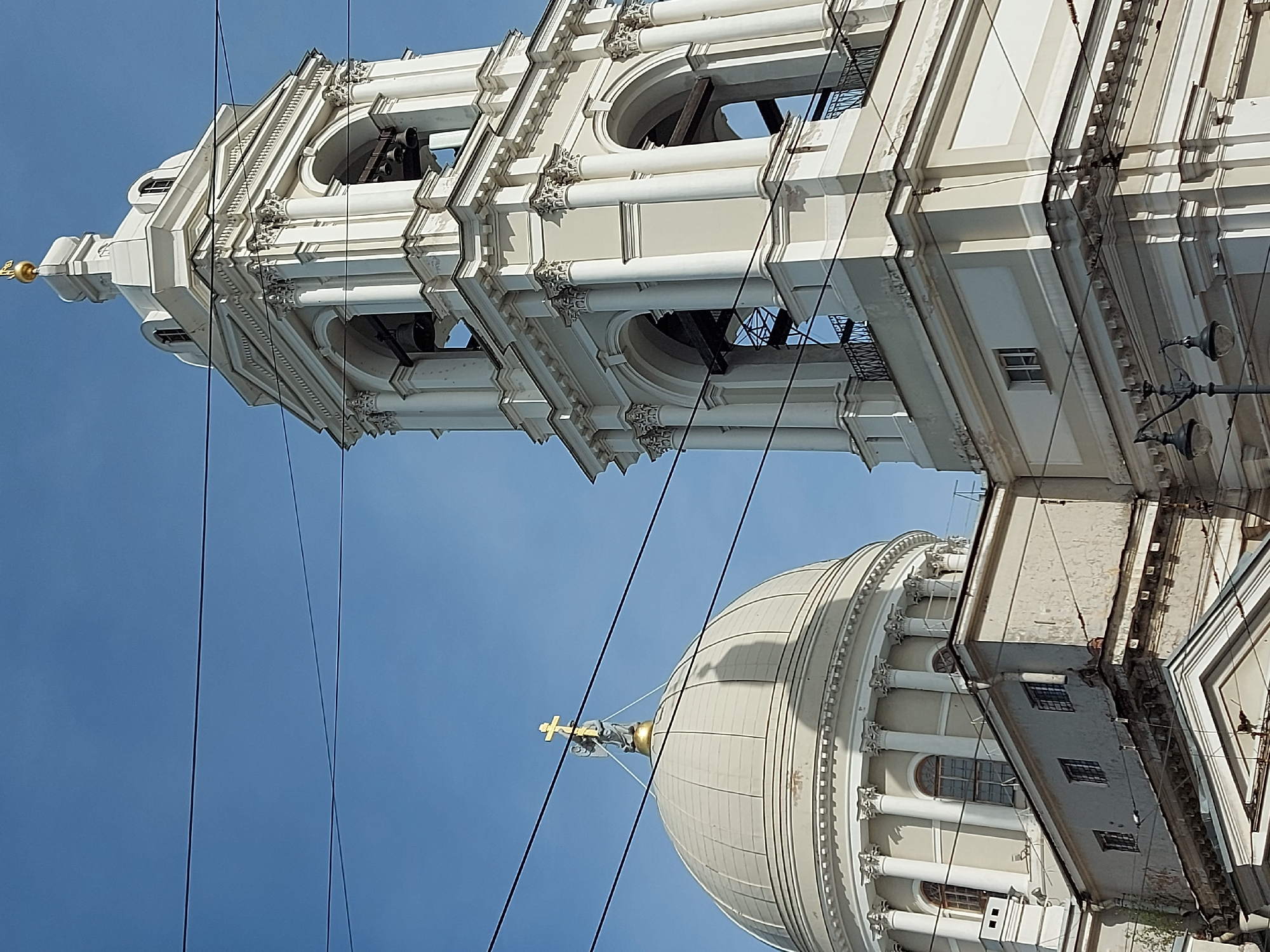 The image depicts the architectural features of the Naval Cathedral of Saint Nicholas in Saint Petersburg, Russia. Visible elements include the bell tower, topped with a golden spire, and a portion of the cathedral's dome, also featuring a golden cross on top. A series of columns and arched openings are visible on both structures. Power lines are running across the view. The background features a clear blue sky.