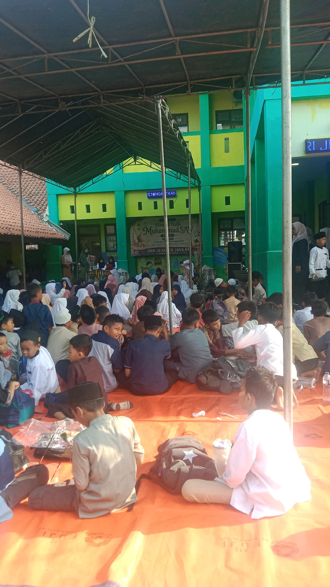 A group of young boys and girls, likely students, are seated on an orange tarp under a shaded outdoor structure at the SDN Jejan Jaya 01 school in Jalenjaya, Indonesia. The children are mostly dressed in traditional Islamic attire, with many wearing white shirts and peci hats, or headscarves. They appear to be engaged in an event, possibly a religious or cultural gathering, as evidenced by a banner in the background that reads "Mau al Nabi Muhammad SAW" and lists the school's name. The building behind them is painted in vibrant green and yellow. Some children are looking towards the front, while others are interacting with their peers or looking down. Objects like backpacks and water bottles are scattered around the tarp, suggesting a casual, communal setting. The overall atmosphere seems peaceful and educational.