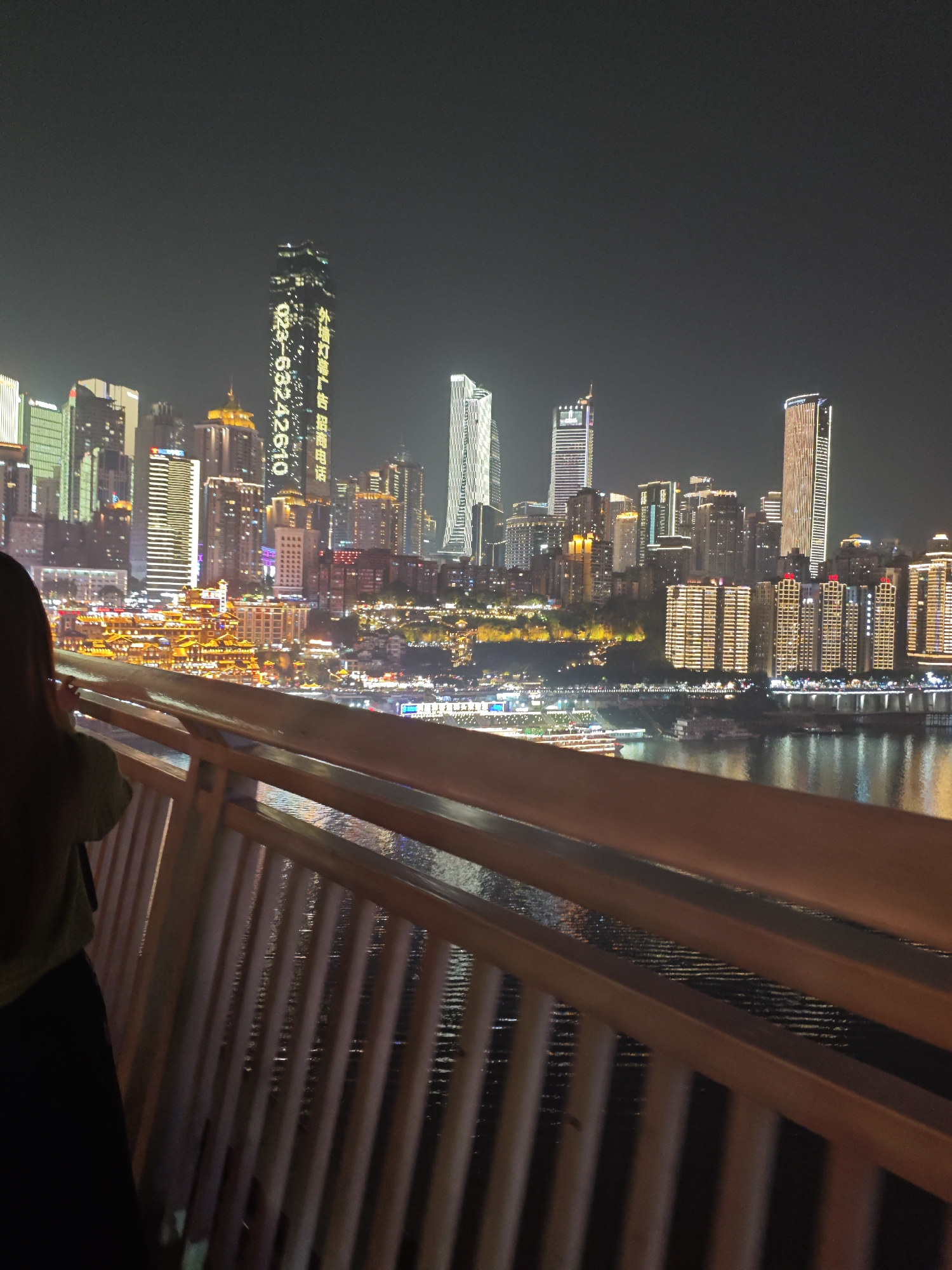 A nighttime view shows a cityscape from an elevated vantage point, likely a bridge or platform. In the foreground, a railing with vertical slats runs horizontally across the frame. Partially visible on the left side of the frame is a person with long hair wearing a dark top, standing near the railing and facing the scene. The background is dominated by a dense urban skyline illuminated with numerous lights. Skyscrapers of varying heights and architectural styles feature prominently, some displaying vertical illuminated signs. Below the skyline, a body of water reflects the city lights. A cluster of traditionally styled buildings with yellow lighting sits near the waterfront. The overall scene is bright with warm hues from the city lights contrasting against the dark night sky and water. This is likely the Yuzhong district of Chongqing, China, given the architectural style of the buildings.