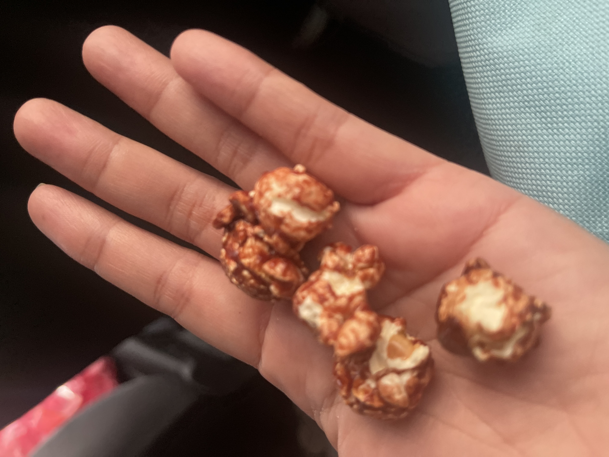 In this close-up, low-angle shot, a person's hand, likely belonging to an adult given the size, is shown open and palm-up, cradling four pieces of caramel-coated popcorn. The popcorn pieces are clustered towards the center and lower part of the palm, with some individual kernels visible. The background is dark and out of focus, with a hint of a light blue textured surface appearing at the bottom right, suggesting it might be fabric. The lighting is dim, creating soft shadows on the hand and popcorn. The overall impression is one of a casual, personal moment, perhaps enjoying a snack. This scene is set in South Tangerang, Indonesia.