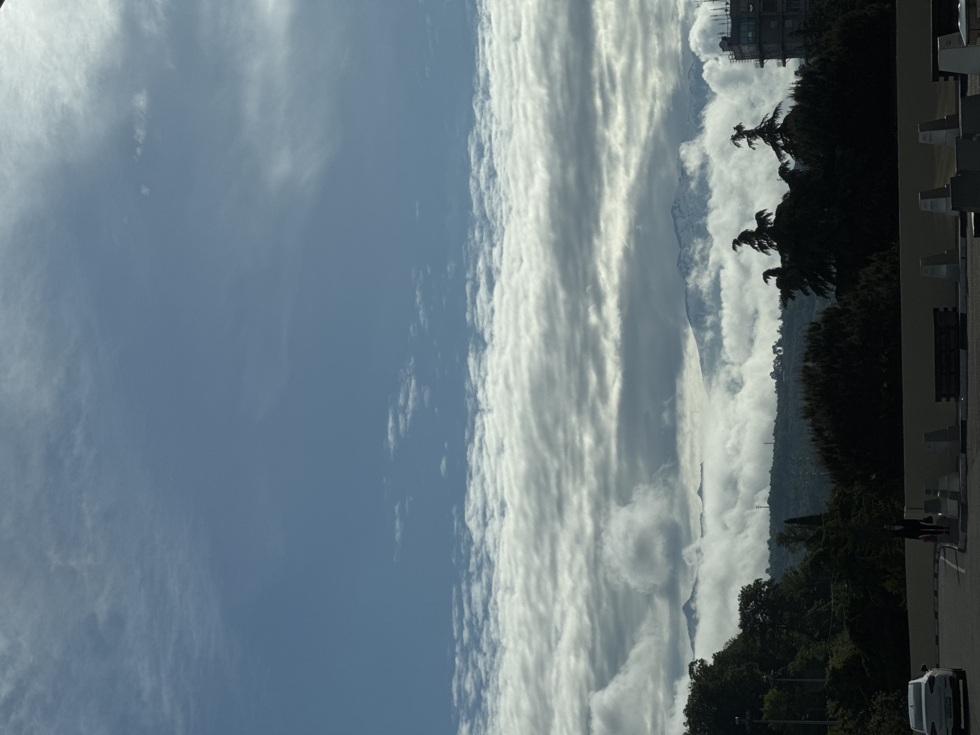 The image shows a view of the sky and landscape in Sochi, Russia. The sky on the left is clear blue with wisps of clouds. To the right, layers of white and grey clouds obscure snow capped mountain peaks. Trees line the edge of the landscape, transitioning to an urban environment on the right, with building and some parked vehicles. A person is walking on the right side of the image.