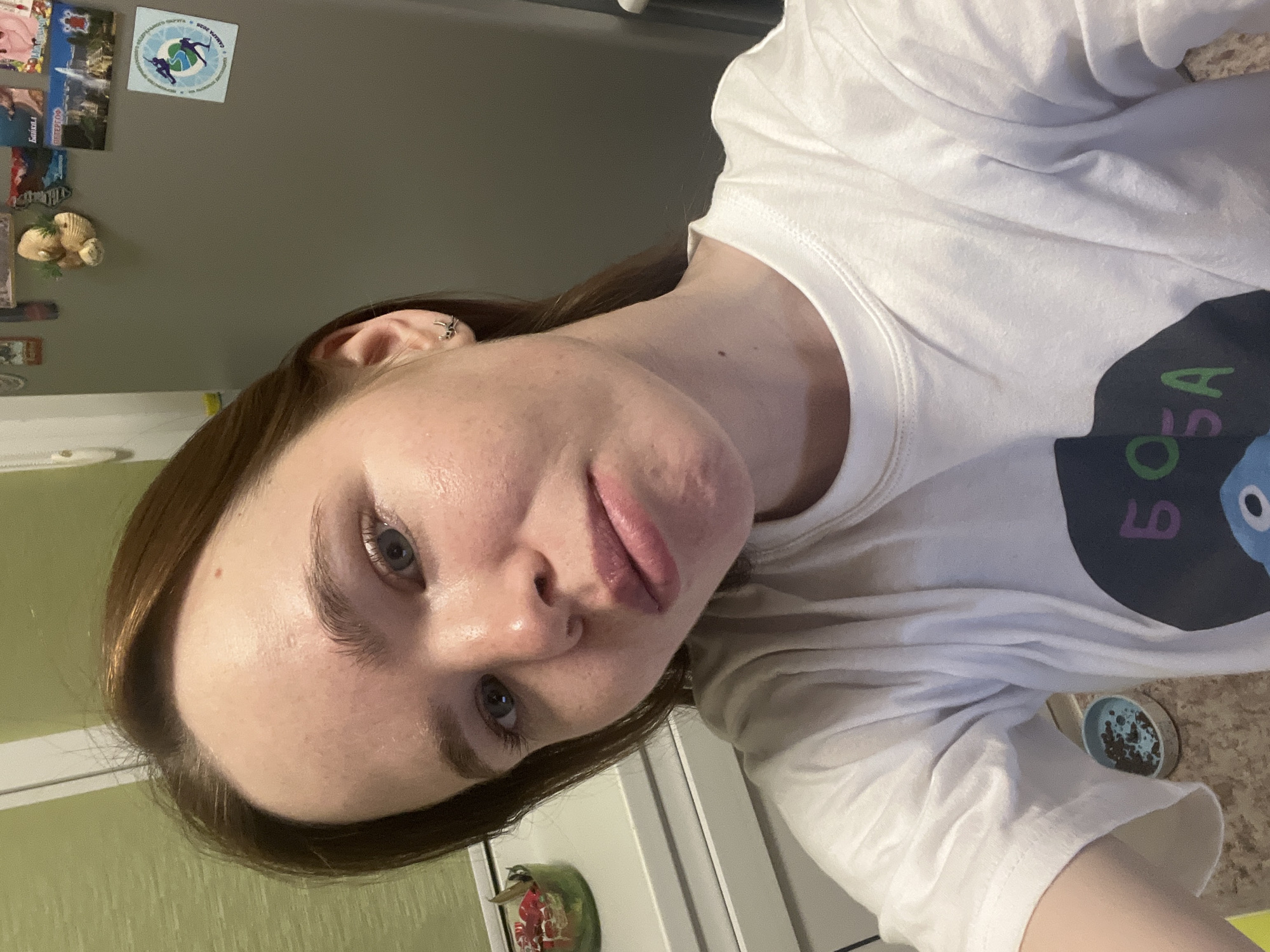 The image is a selfie of a young woman, taken indoors in a home setting, likely in Chaikovsky, Russia, given the provided context and visible Cyrillic text. The photo is rotated, showing the woman's head tilted.
**Subjects:** The central subject is a young woman with fair skin, brown hair, and blue eyes, looking directly at the camera. She has an ear piercing (a small silver hoop or stud) in her right ear.
**Objects:** She is wearing a white short-sleeved t-shirt with a dark blue graphic on the right side of her chest, featuring a stylized character and green Cyrillic text. The background features a gray wall, possibly a refrigerator door, adorned with several magnets. These magnets include a circular one with a design resembling a globe or world map, and small figures, along with visible Cyrillic text including "СБОРНАЯ РОССИИ" (Russian National Team), "КАНАДА" (Canada), and "2019". Other rectangular magnets show landscapes or cities, with one clearly reading "БАЙКАЛ" (Baikal) and another "ПИТЕР" (Piter/St. Petersburg). A small decorative object resembling seashells is also attached. Below this, a light brown patterned countertop is visible, on which a small, light blue round bowl containing dark, granular material (possibly coffee grounds or soil) is placed.
**Activity/Event:** The woman is posing for a self-portrait.
**Notable details:** Her expression is neutral. Her skin appears slightly shiny on her forehead and cheeks. Her lips are slightly parted. The visible Cyrillic text on the magnets and her t-shirt, alongside the location context, strongly indicates a Russian locale.
**Visible text:** On the t-shirt, the Cyrillic text "БОБА" is visible. On the magnets, "БАЙКАЛ", "ПИТЕР", "СБОРНАЯ РОССИИ", "КАНАДА", and "2019" are discernible.