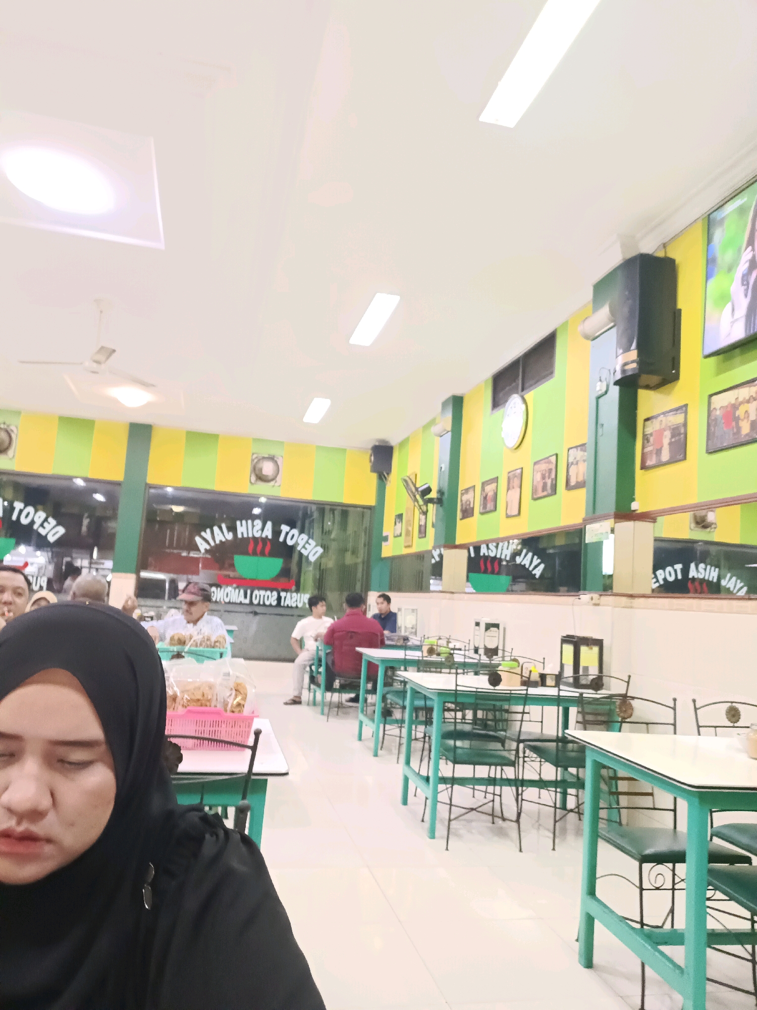 The image shows the interior of a restaurant in Dapur, Indonesia, with a woman in a black hijab in the foreground. The restaurant features green and yellow striped walls and green tables with white tops and green chairs. There are framed pictures on the walls and a television screen displaying an image of a woman. In the background, several people are seated at tables. Signs with text are visible on the glass windows and walls. The ceiling has fluorescent lighting and a ventilation fan.