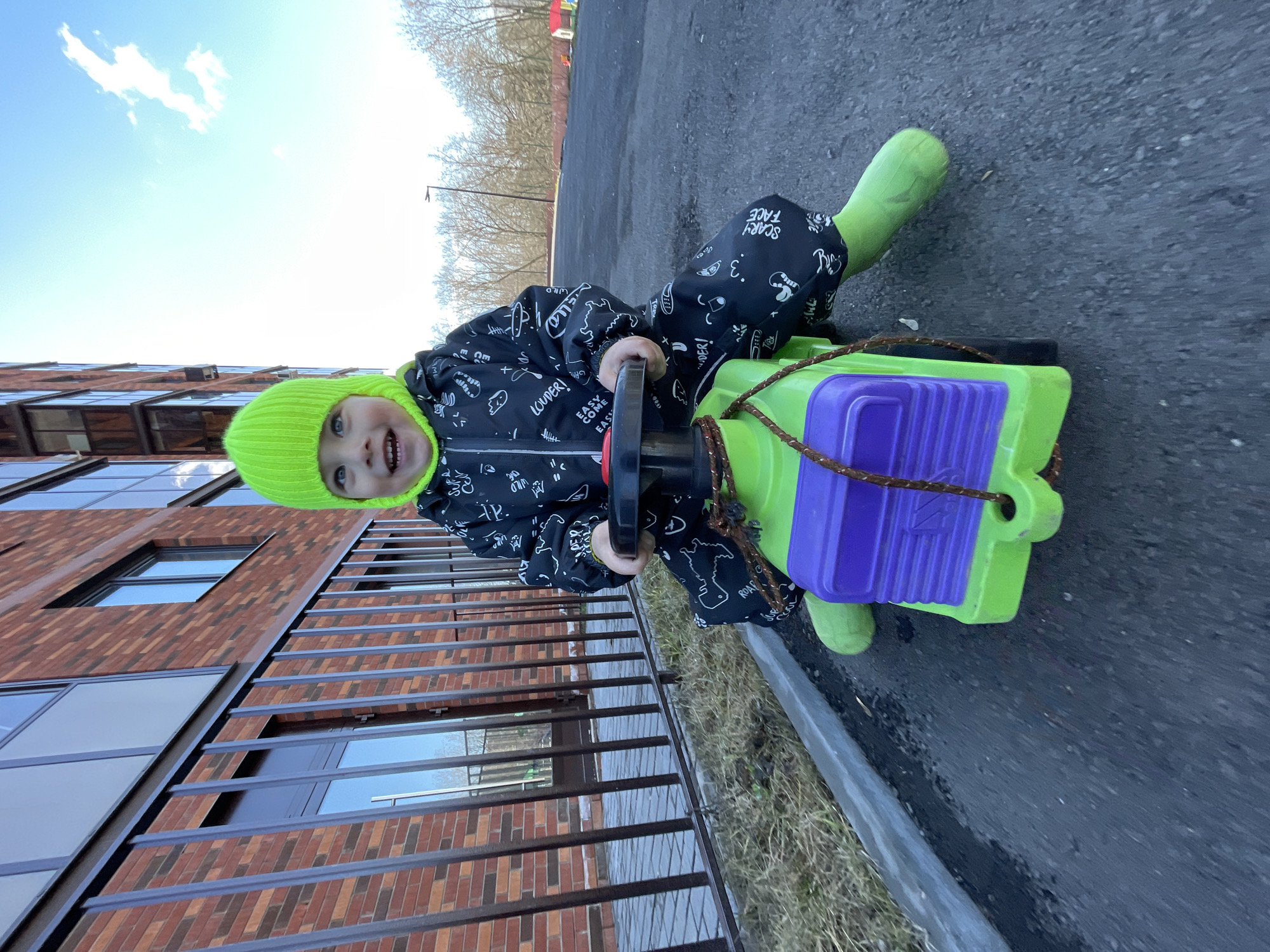 The image captures a young child outdoors, rotated 90 degrees to the left.
**Subjects:** The primary subject is a smiling child, likely a toddler, wearing a bright neon green knitted beanie hat and matching neon green rubber boots. They are dressed in a black full-body suit or jumpsuit adorned with white patterns and text. In the background, a modern brick building with large rectangular windows is visible on the left, and bare trees are present in the upper right.
**Objects:** The child is actively engaged with a brightly colored pull-along toy. The toy is predominantly neon green with a purple section on top, featuring a black handle and a brown rope for pulling. The ground beneath the child and toy is a dark asphalt or paved surface, with some patches of dry grass along a curb next to the building. The sky is bright blue with a few white clouds.
**Activity or event taking place:** The child is sitting on or beside their pull-along toy, looking directly at the camera with a wide, joyful smile, suggesting an active playtime moment.
**Notable details:** The scene depicts a bright, clear day, likely late winter or early spring given the bare trees. The child's warm attire (hat, jumpsuit, boots) also suggests cooler weather. The modern architectural style of the building provides a distinct urban or suburban setting. The child's expression conveys happiness and excitement. The location context is Izhevsk, Russia.
**Visible text:** Text visible on the child's jumpsuit includes "SCARY FACE", "LOUDER!", "EASY COME EASY", "ROAR", "WILD", and the number "3".
