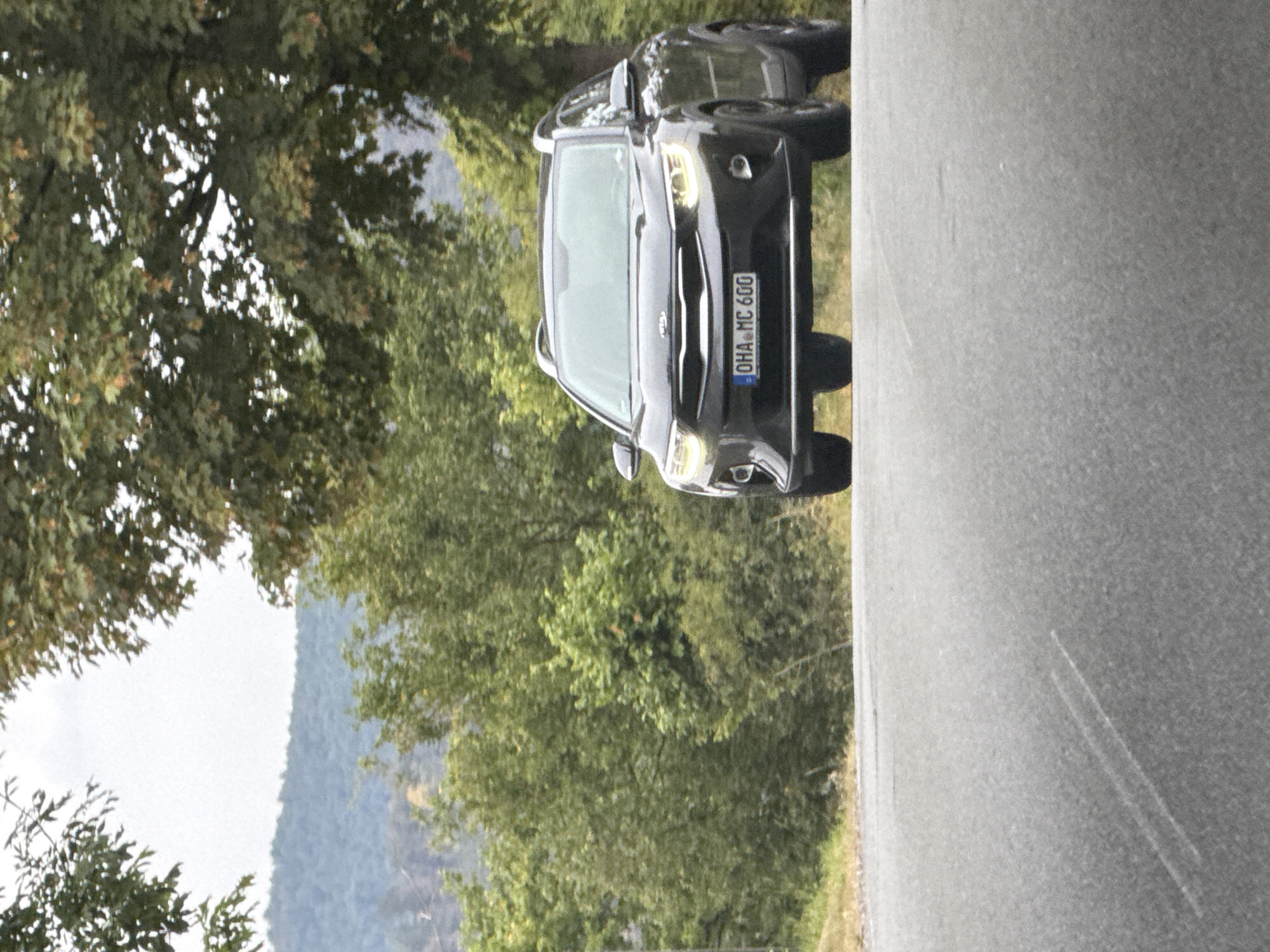 An eye-level, medium shot captures a dark-colored Kia Stonic SUV driving on an asphalt road. The car is facing the viewer and is positioned slightly to the right of the frame's center. The car's license plate reads "OHA MC 600". Behind the car, a lush green forest covers rolling hills, and a cloudy sky is visible in the distance. The road in the foreground is marked with white lines. The scene appears to be located in Bad Lauterberg im Harz, Germany.