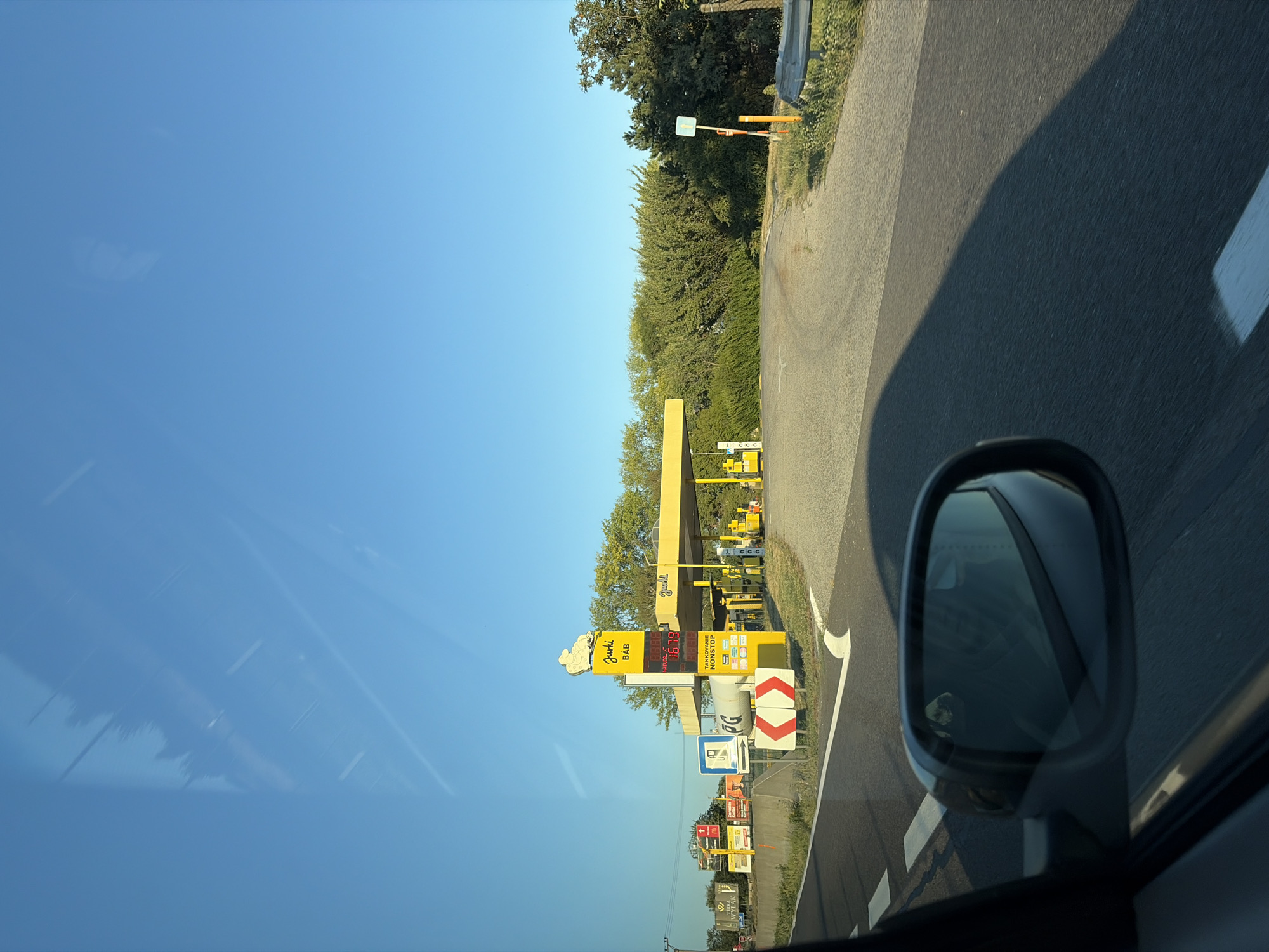 The image shows a view from a car traveling along a road in the vicinity of Báb, Slovakia. On the right side of the road, a yellow gas station with a prominent sign featuring the logo "Jurki BAB" is visible. The gas station has a canopy covering multiple fuel pumps. A digital price display shows fuel prices, with one reading "1.679" and another "1.63". Adjacent to the gas station are various advertisements and signs, including one with a blue symbol and directional arrows, and another with red and white chevron markings indicating a curve. The surrounding environment consists of trees, vegetation, and a clear blue sky. The car's side mirror is also visible in the foreground, reflecting parts of the interior.