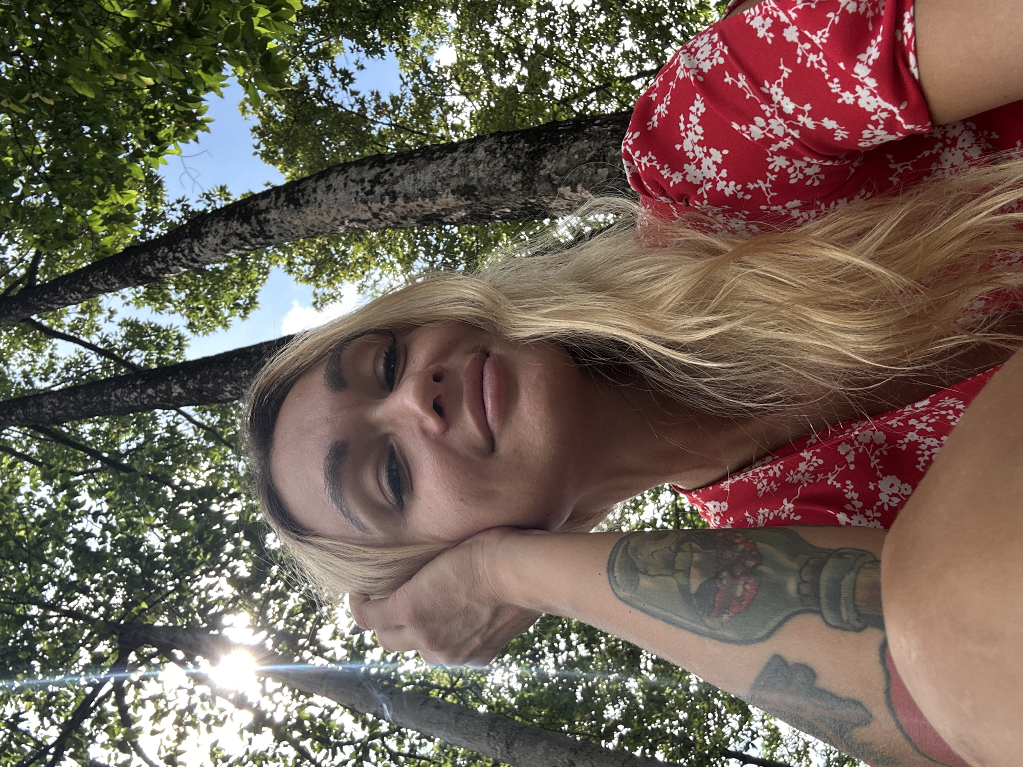 A blonde woman in a red floral dress rests her head on her tattooed left arm. Trees with green leaves are visible behind her, with sunlight filtering through the foliage. The sky is partially visible through the trees. The location is most likely outdoors in a park or wooded area, potentially within the province of Đồng Nai, Vietnam.