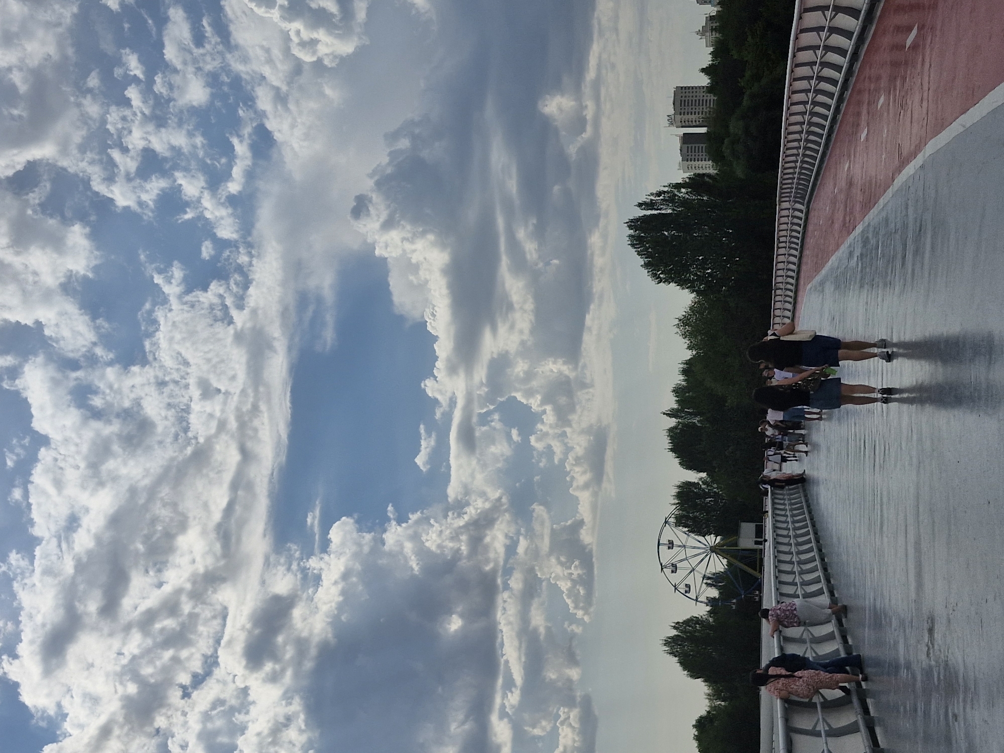 The image depicts a wide pedestrian bridge with a red lane for cycling or running. Several people are walking on the bridge. In the background, there is a Ferris wheel, trees, and some buildings, suggesting an urban park or recreational area. The sky is filled with dramatic, white and gray clouds. The image appears to be taken in Astana, Kazakhstan, a city known for its modern architecture and public spaces, which often include elevated walkways and recreational facilities like this bridge.