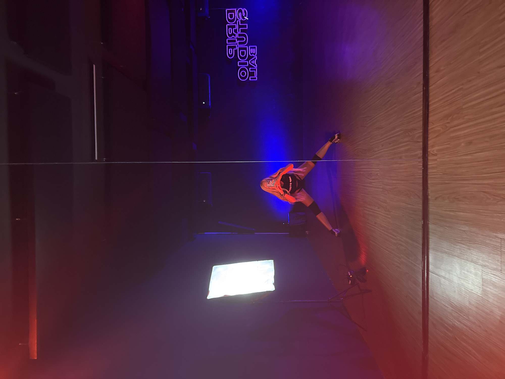 The image captures the interior of a space identified as "Bali Drip Studio," in North Kuta, Indonesia, with the studio's name visible in reverse lettering on a dark wall. A woman, seemingly in a dancer's or athletic outfit consisting of an orange top and black shorts, stands in the studio. A large, bright rectangular light, probably a photographic softbox, illuminates the space, positioned on a stand. The floor is a dark material, transitioning into a lighter woodgrain pattern on one side. The walls are dark, with what appears to be a mirrored wall on the left and another dark wall behind the woman. The overall lighting of the scene creates vibrant colors, with purplish and reddish hues.