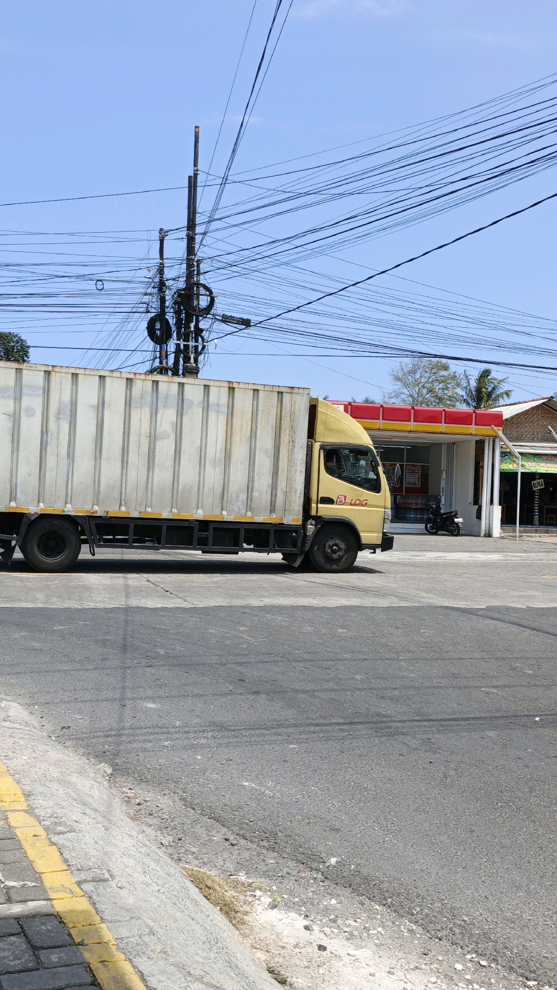 A yellow box truck with a white cargo container is parked on a street in Cibenda, Indonesia. The truck is positioned on the right side of the frame, with its front facing the right. The truck has a logo that reads "B LOG" on its side. In the background, a building with a red and yellow awning appears to be a shop or convenience store. A motorcycle is parked in front of the building. The street is paved with asphalt, and there is a yellow line marking the edge of the road and a sidewalk. Overhead, a complex network of electrical wires is suspended from poles. The sky is clear and blue.