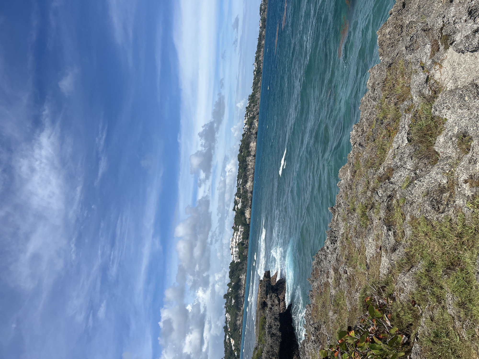 The image depicts a coastal scene near Le Gosier, France. The view is taken from a cliff overlooking the ocean. The water is turquoise-blue, with waves crashing against the rocky shoreline. In the distance, there is a coastline dotted with buildings, suggesting a residential or resort area. The sky is partly cloudy with streaks of blue visible. The cliff face is covered with vegetation, including grass and shrubs.