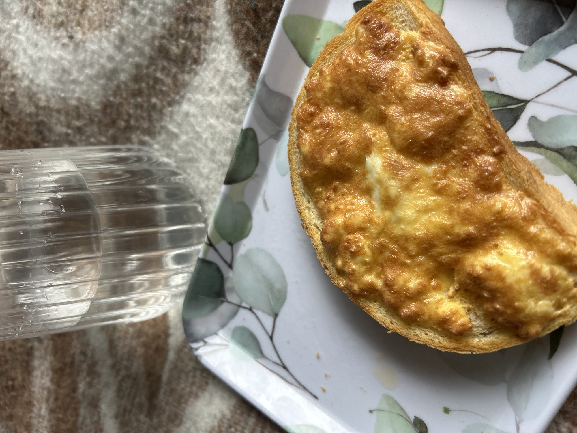 A slice of cheese toast is on a floral patterned tray. A partially-filled glass of water is to the left of the tray. A brown and white blanket is to the left of the water. The location in Burgas, Bulgaria is not discernible from the image.