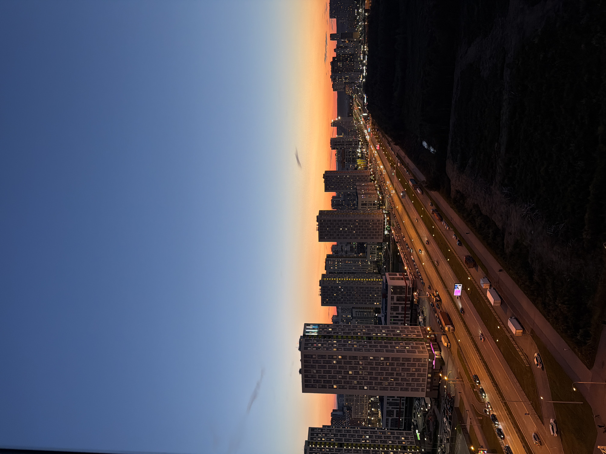 The image captures a twilight view of a city, characterized by a gradient of colors in the sky from deep blue at the top to vibrant orange and pink near the horizon. Dominating the foreground are several tall, modern apartment buildings with numerous windows, many of which are illuminated. A multi-lane highway runs through the scene, with traffic visible in the form of headlights and taillights of cars and trucks, indicating active movement. Streetlights line the highway, casting a warm glow. To the right of the highway, a dark, densely wooded area is visible, contrasting with the illuminated urban landscape. The overall impression is that of a sprawling residential district at dusk, with the city lights beginning to dominate as natural light fades. Without additional information, the specific location within Yekaterinburg, Russia, cannot be determined from the image alone.
