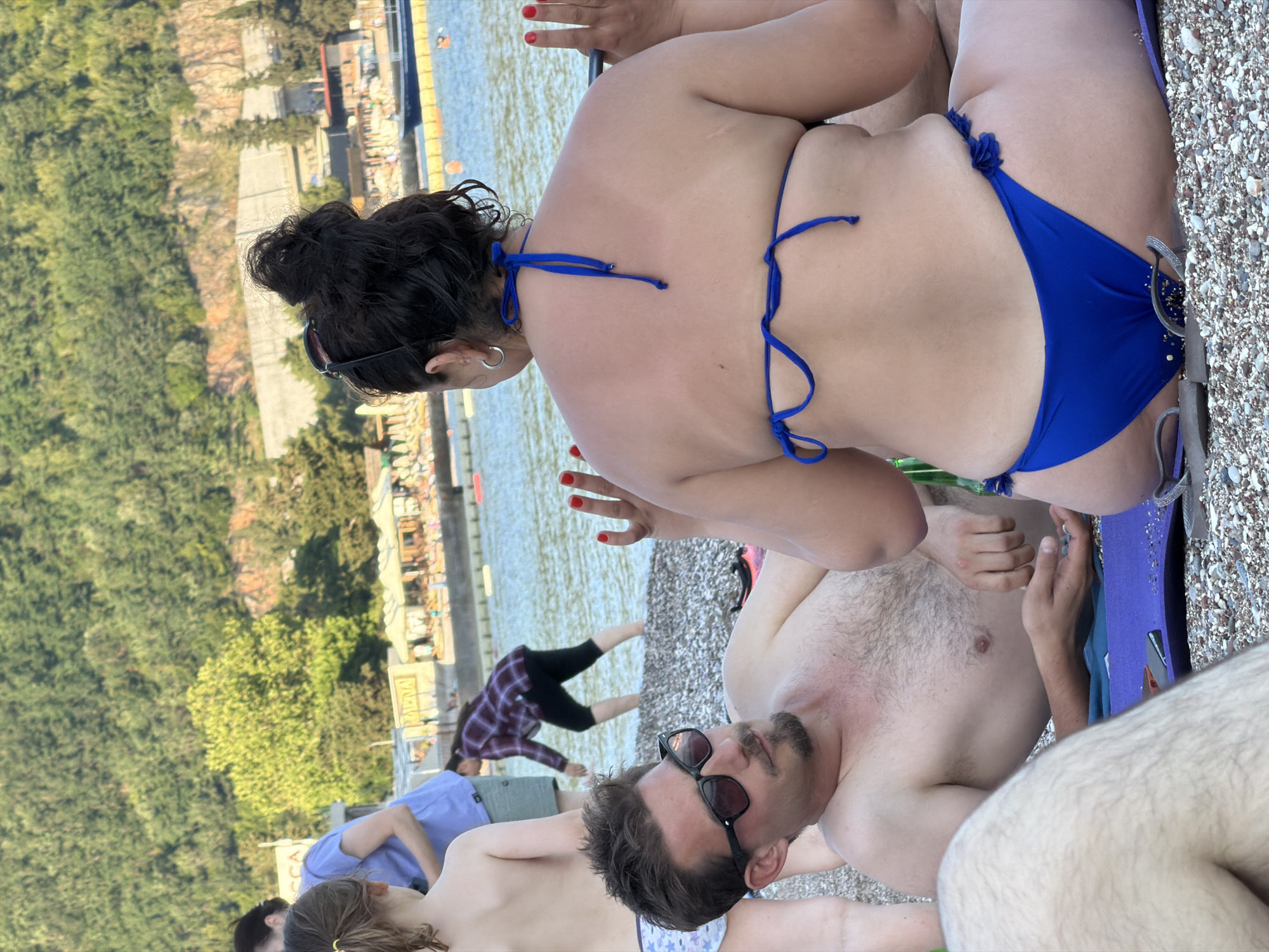 The image shows a beach scene in Budva, Montenegro. A woman with dark hair in a bun, wearing a blue bikini, is the central focus. She is sitting on a purple beach chair. To her side, a man with dark hair and sunglasses is reclining, shirtless. Another person, partly visible, is lying near him. A person in a plaid shirt is visible in the background near the water. The beach is composed of small pebbles. In the background, there are buildings, trees, and water, characteristic of a coastal town like Budva.