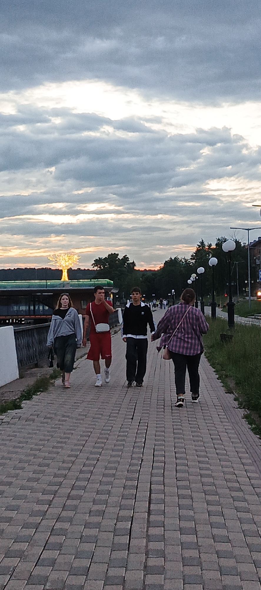 Four individuals are walking on a brick-paved promenade in Votkinsk, Russia, during twilight. The sky is filled with dramatic, cloud-filled patterns with hints of orange and yellow from the setting sun. To the left, a luminous, tree-shaped sculpture is visible, and beyond it, a body of water and trees can be seen. The promenade is lined with lampposts, some with spherical lights, and a grassy area is to the right. The people are walking away from the camera, dressed in casual attire. The person on the far left is wearing a grey hooded jacket and dark pants. Next to them is a person in a red t-shirt and red shorts, carrying a light-colored crossbody bag. The third person is in a black long-sleeved shirt and dark pants. The person on the far right is wearing a purple and pink plaid shirt and dark pants.
