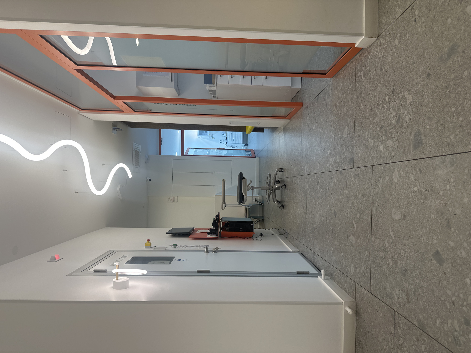 The image captures an interior view, likely a hallway or corridor, within a modern medical or laboratory facility. The space is characterized by a clean, sterile aesthetic with white walls and a gray, speckled tile floor. On the left, a white, cleanroom-style door features a small window and industrial hardware. Above it, a modern, wavy light fixture adds an artistic touch. Further down the hall, a glass-enclosed room with an orange frame is labeled "STERILISATION," hinting at the facility's purpose. Inside, glimpses of medical equipment and storage can be seen. The hallway contains an examination chair with wheels and medical equipment, suggesting this is a functional space for patient care or research. The location is unknown.