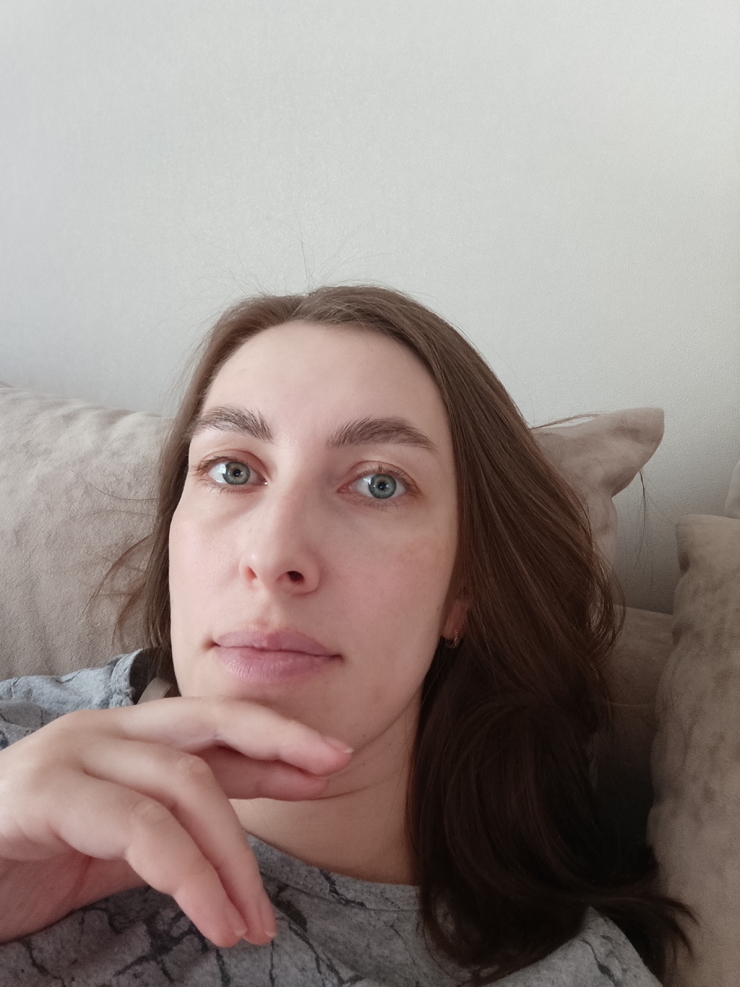 The image is a close-up, slightly upward-angled self-portrait of a young adult woman. She has long, straight brown hair, parted on the left side, which frames her face. Her striking blue-green eyes are wide open and gaze directly at the viewer with a calm, neutral expression. Her eyebrows are full and naturally defined. She has a fair complexion and appears to be wearing minimal or no makeup.
Her right hand is positioned with fingers gently curled, resting beneath her chin. She is wearing a light grey short-sleeved top with a subtle, darker grey abstract pattern.
The background suggests an indoor setting, likely a home environment. A plain, light off-white or light grey wall is visible behind her head and to her left. To her right, a portion of a light beige or tan upholstered sofa or cushion is present, indicating she is either sitting or reclining against it. The scene is illuminated by soft, natural light, suggesting it is daytime. There is no visible text in the image.