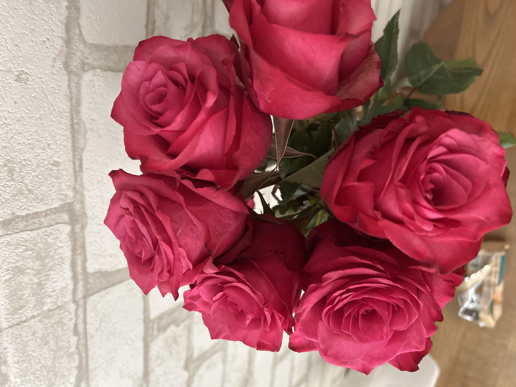 A bouquet of vibrant pink roses is displayed in front of a textured white brick wall. The roses, with their tightly furled petals, dominate the foreground of the image. The lighting casts subtle shadows from the roses onto the wall, adding depth to the scene. The lower portion of the image shows a wooden surface and a glimpse of green leaves. The overall impression is one of natural beauty and simple elegance. The location is in Ivanivka, Ukraine.