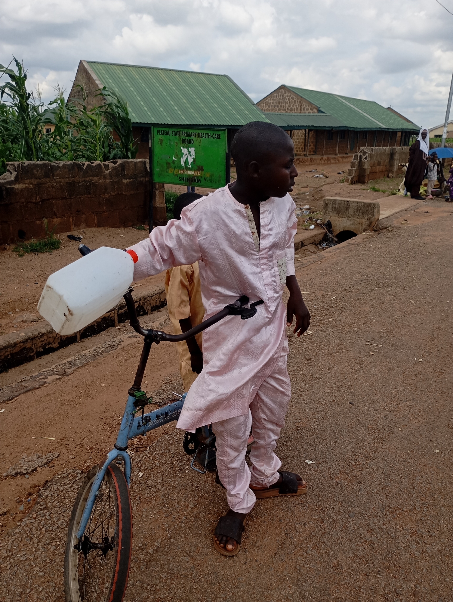 A boy in a light pink traditional outfit stands beside a blue bicycle in Shendam, Nigeria. He holds a white plastic jerrycan in his right hand, which is attached to the bicycle's handlebars. The boy wears dark sandals. In the background, there are several buildings with green corrugated iron roofs, a brick wall, and a green sign that reads "PLATEAU STATE PRIMARY HEALTH-CARE BOARD, SHENDAM." A second boy, wearing a yellow outfit, is partially visible behind the first boy. Other individuals and a blue umbrella are seen further in the background on a dirt road. The sky is overcast with clouds.