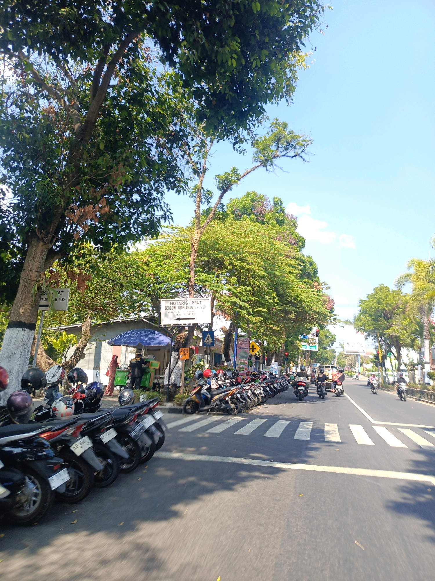 The image depicts a street scene in Tambran, Indonesia, on a sunny day. The street is lined with parked motorcycles on the left side of the frame, with a pedestrian crossing marked by white stripes in the foreground. Lush green trees with dense foliage overhang the street and sidewalk. On the left sidewalk, a small food stall is visible under a blue umbrella, with several people standing around it. To the right of the food stall, there are signs, including one that reads "NOTARIS.PPAT SETO DWI KURNIAWAN, S.H., M.Kn" with an arrow pointing to the right. Further down the street, traffic is moderate, with several motorcycles and a few cars visible. The sky is clear blue with a few scattered clouds. The overall impression is of a typical urban street in Indonesia, with vibrant greenery and bustling activity.