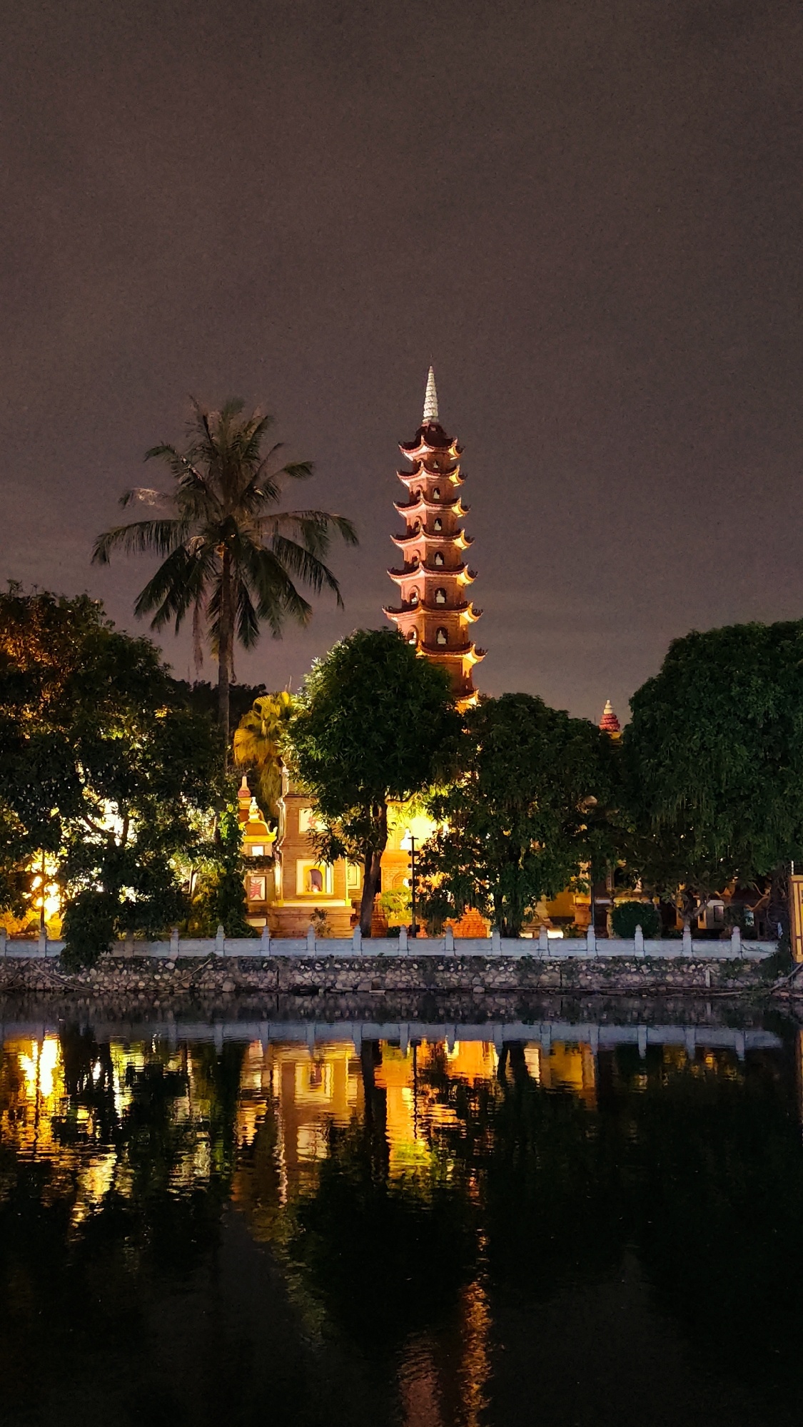 The image captures a nighttime view of the Tran Quoc Pagoda in Hanoi, Vietnam. The pagoda, a tall, multi-tiered structure with an orange-red facade and a white spire, is illuminated against a dark, cloudy sky. It is situated behind a stone wall and a row of lush green trees, some of which are also lit from below. A palm tree stands to the left of the pagoda. In the foreground, a dark body of water reflects the lights of the pagoda and surrounding trees, creating an impressionistic mirrored image. The scene is serene and atmospheric, highlighting the architectural beauty of the pagoda and its tranquil setting.
