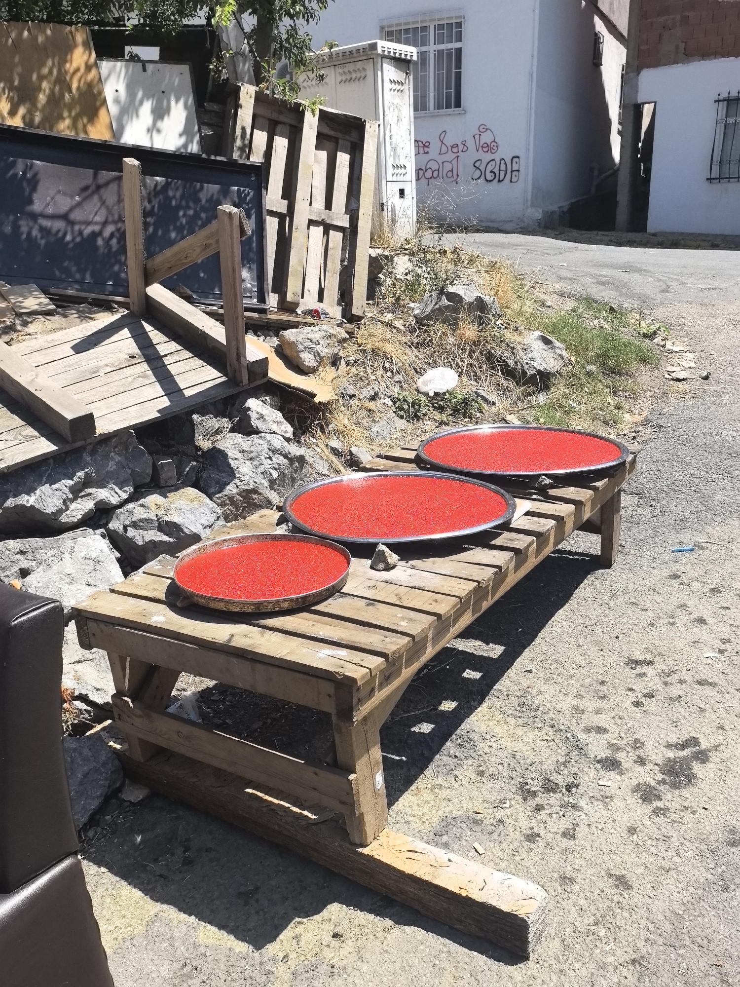 In Ataşehir, Turkey, three large, shallow metal trays are filled with a bright red, granular substance, possibly paprika or chili flakes, and are placed on a rustic wooden table. The table is constructed from rough-hewn planks and stands on uneven ground. Behind the table, large gray rocks and dried vegetation are visible. In the background, a white building with barred windows and graffiti on its facade is seen, along with stacks of wooden pallets and other discarded materials. The scene appears to be outdoors, under bright sunlight, suggesting it may be a street or an open yard area.