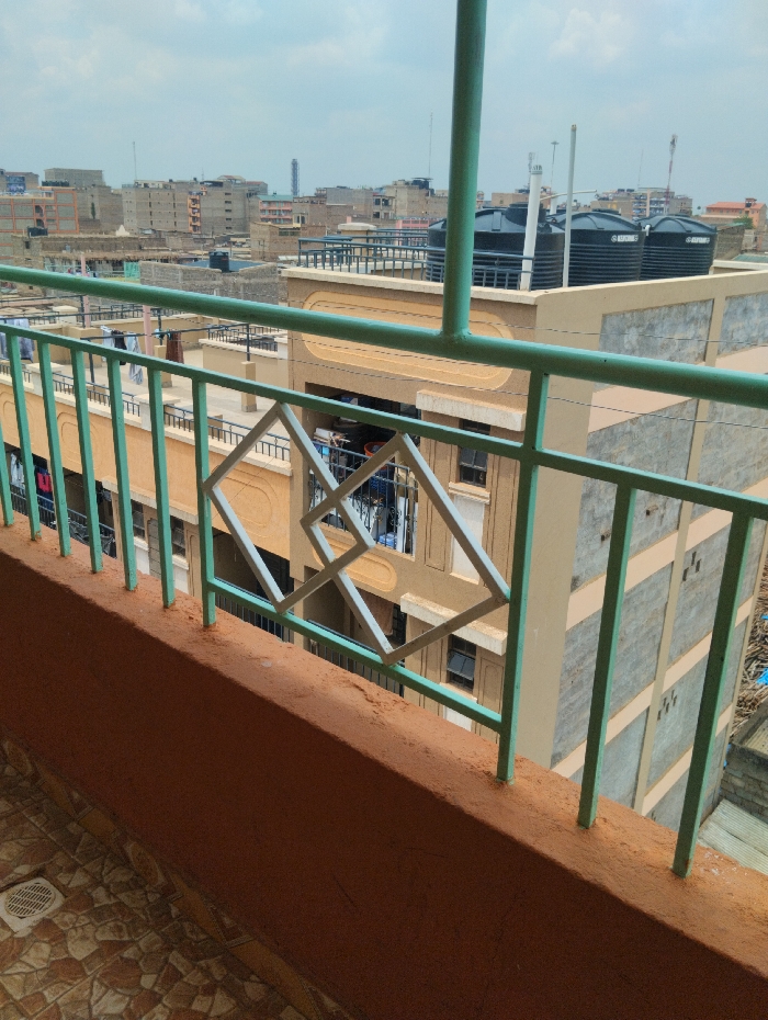 The image depicts an elevated view from a balcony overlooking a densely built urban environment in Witeithie ward, Kenya.
In the immediate foreground, a tiled floor with brown and beige geometric patterns is visible on the left, including a white circular drain cover. A reddish-brown concrete parapet wall spans the lower half of the frame, topped by a mint green metal railing. The railing consists of vertical bars interspersed with a decorative motif of interlocking square-diamond shapes. The paint on the railing shows signs of light wear.
Beyond the railing, the midground is dominated by multiple multi-story residential buildings. An adjacent building directly across from the viewpoint is tan with brown accents, displaying various articles of laundry hanging from its balconies. To the right, another building, lighter in color, exhibits visible concrete structural elements. On the rooftop of a prominent building in the midground, three large, black plastic water tanks, labeled "SAFI WATER TANKS," are clearly visible, accompanied by several white poles and antennae.
The background presents a sprawling urban landscape characterized by numerous multi-story structures of varying heights and designs, extending towards a hazy horizon under a light blue, cloudy sky. A tall, slender industrial-style structure is discernible on the distant left horizon. Multiple communication towers or antennae are visible atop distant buildings. No individuals are visible in the scene, but the presence of laundry suggests human habitation.