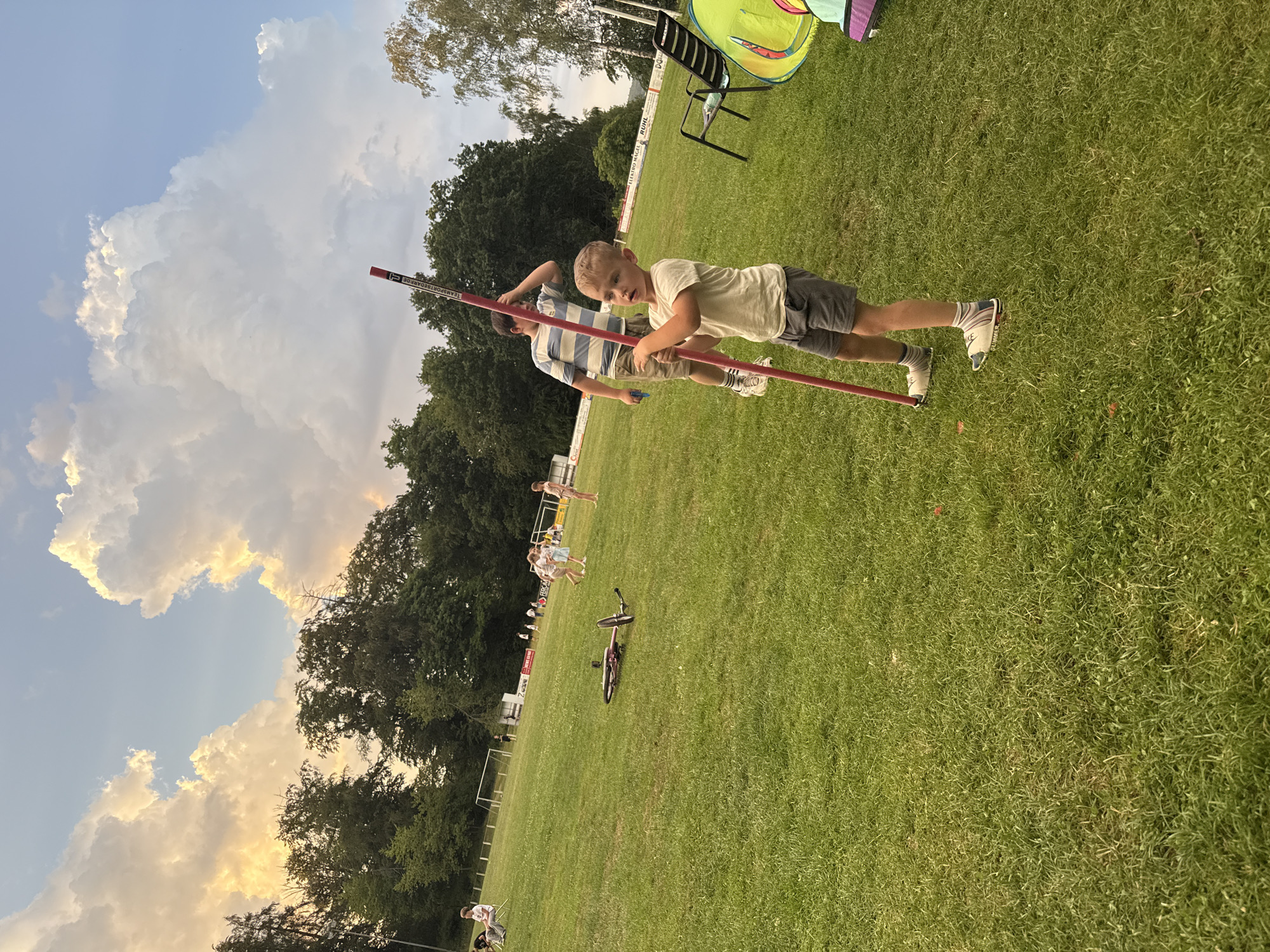 In Lumda, Germany, on a grassy field, a young boy stands in the foreground, holding a red pole with white markings. He wears a light-colored t-shirt, grey shorts, striped socks, and white and colored sandals. Behind him, an older boy in a striped t-shirt and beige shorts stands with his hand on his forehead. The background shows a treeline, a soccer field with goals, several people walking and standing, and a bicycle lying on the grass. A chair and a colorful tent are also visible. The sky is filled with dramatic clouds.