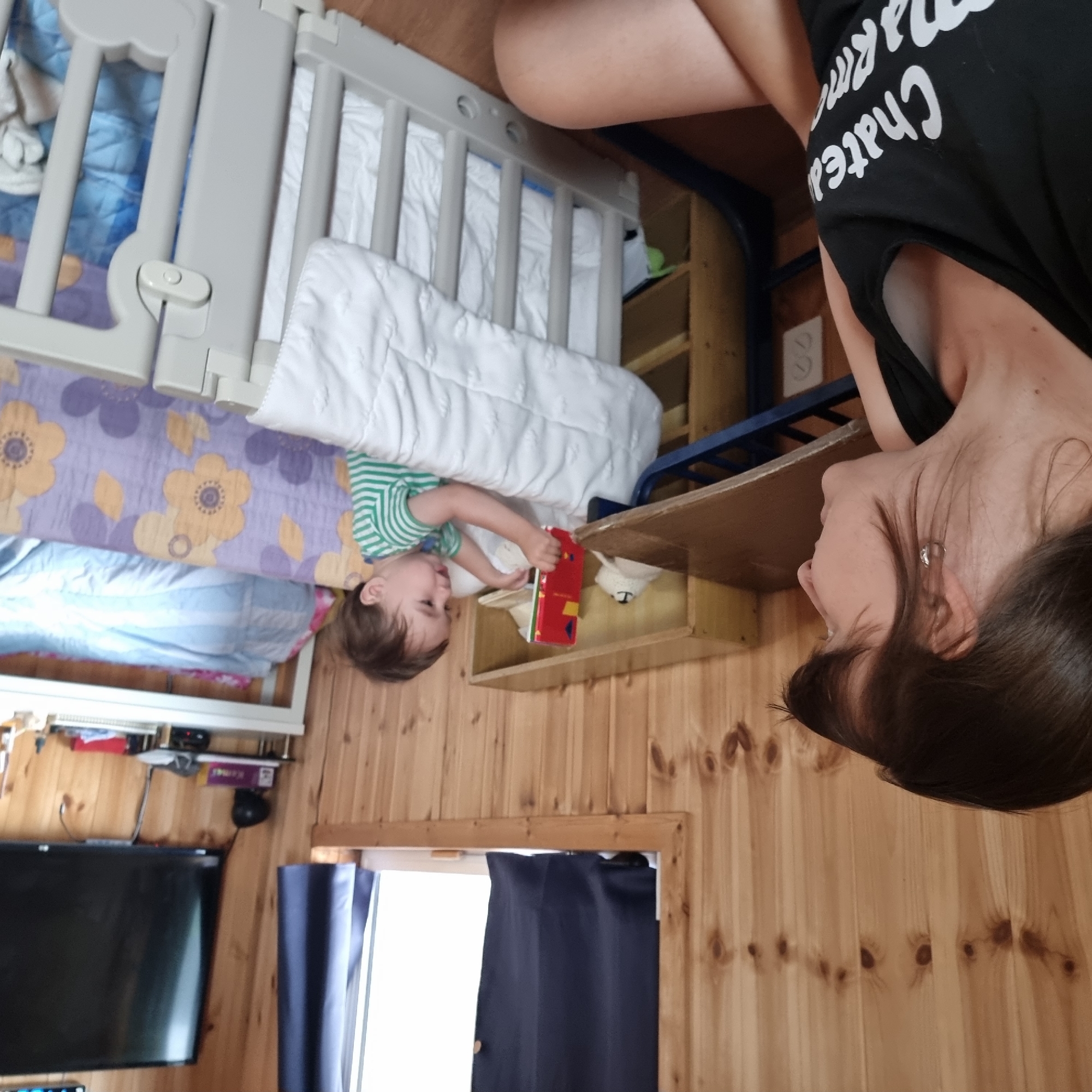 The image shows a woman in the foreground with dark hair, wearing a black tank top with white text. She is looking to her right, in profile. To her left and slightly behind her, a young child wearing a green and white striped shirt is sitting in a crib and holding a red object. The crib has white bedding and a patterned bumper with yellow and purple flowers. The background consists of wooden walls. To the right of the crib, a black television is mounted on the wall above a white shelf with various items. The scene appears to be indoors, possibly a bedroom or play area. There is no specific indication of the location being in Dunnae-myeon, South Korea.