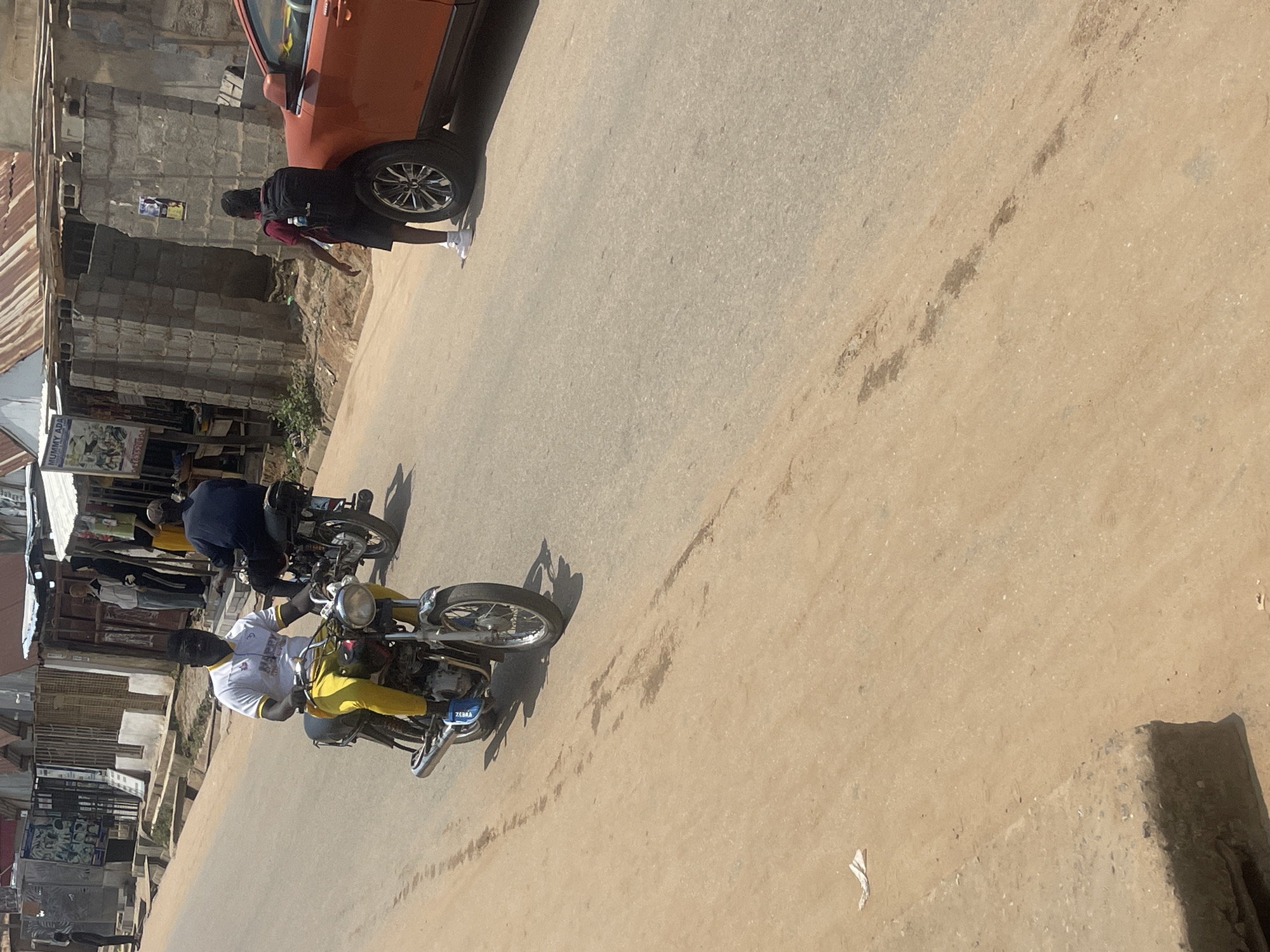 This image depicts a street scene in Abuja, Nigeria, under a bright sky. In the foreground, a man wearing a white shirt with a logo and yellow pants rides a motorcycle. Behind him, another person on a motorcycle is partially visible. Further down the street, a person wearing a maroon shirt and shorts, with a black backpack, stands next to an orange car. The environment includes buildings with corrugated metal roofs and stalls lining the street. The road surface is a mix of asphalt and dirt.